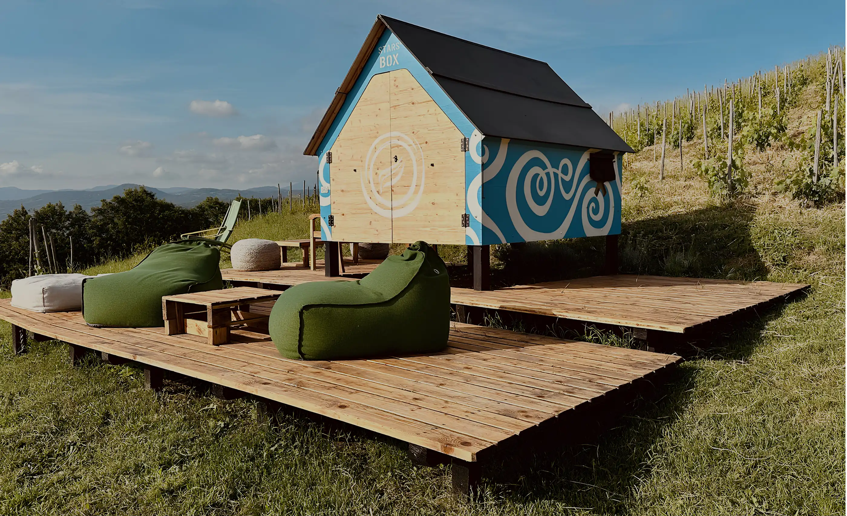 A small blue and timber hut on stilts marked Stars Box, with green bean bags on a wooden deck beside a vine slope