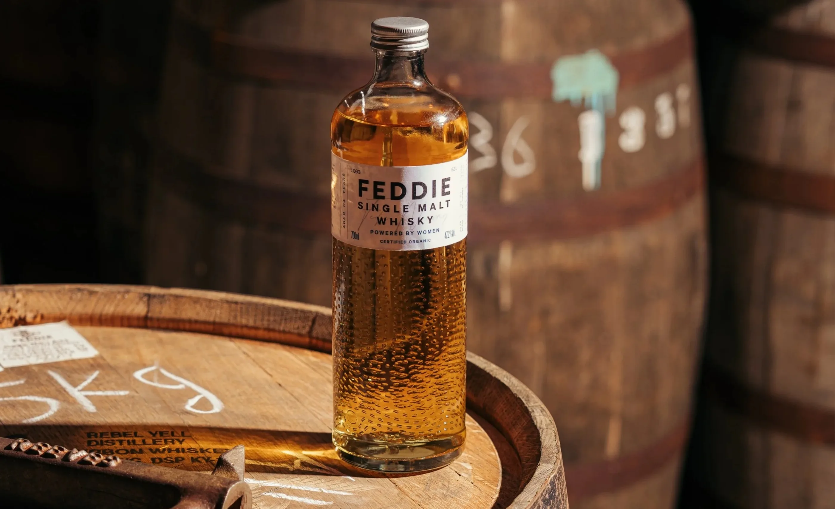 A bottle of Feddie single malt whisky standing on a chalk-marked barrel head, with more barrels behind