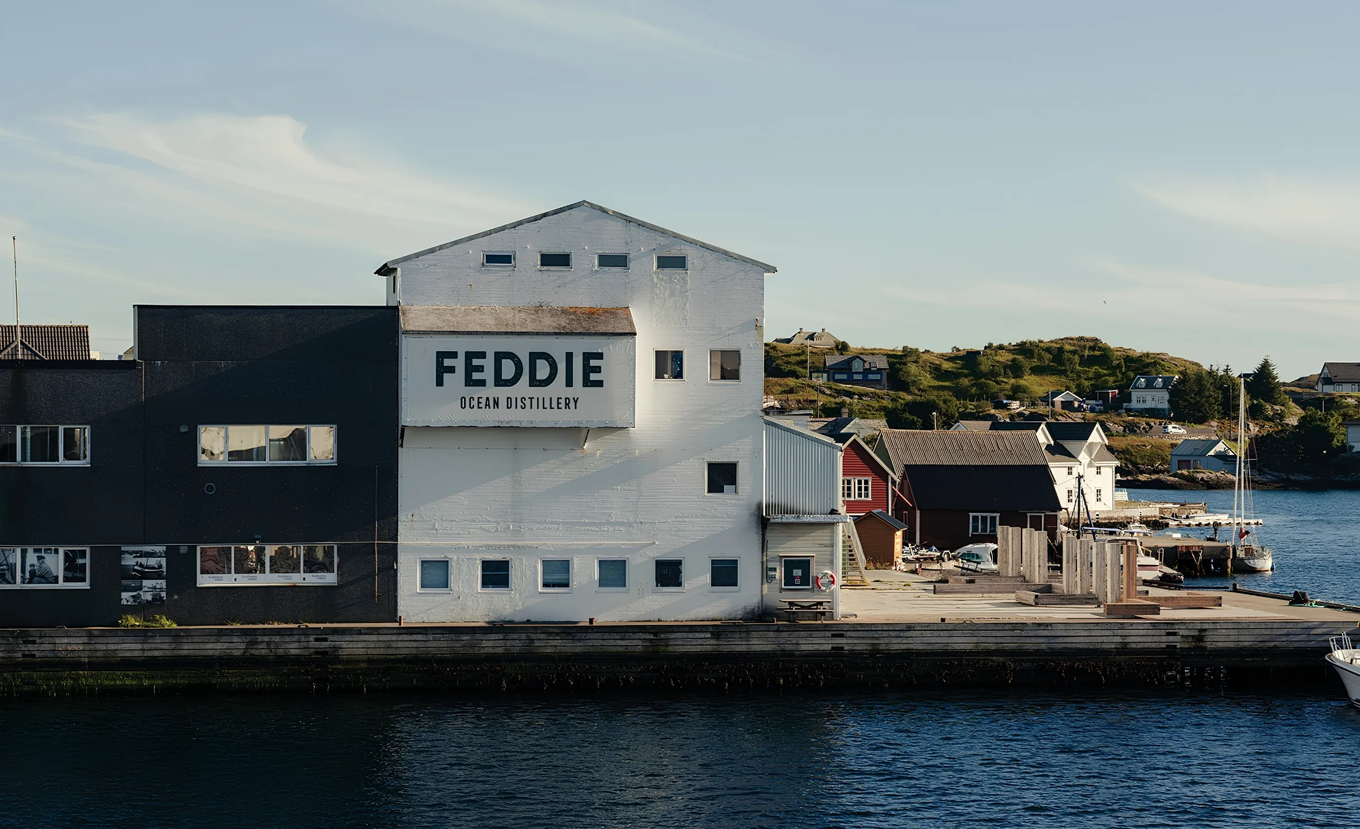White waterfront building with a Feddie Ocean Distillery sign, and small houses and a sailboat on the far shore