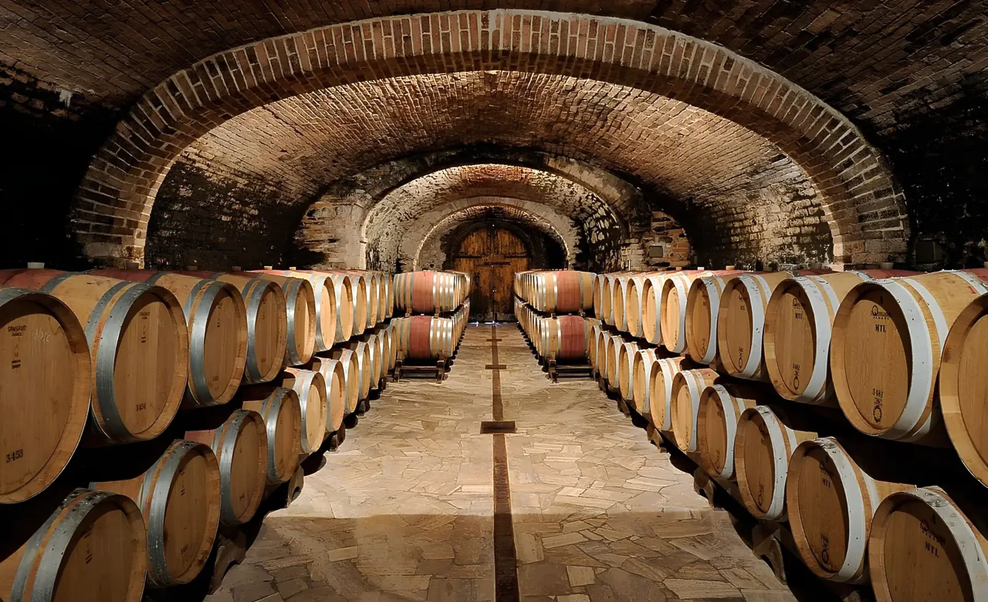 Oak barrels stacked two high on both sides of a brick-vaulted cellar, with a stone-paved aisle leading to a wooden door
