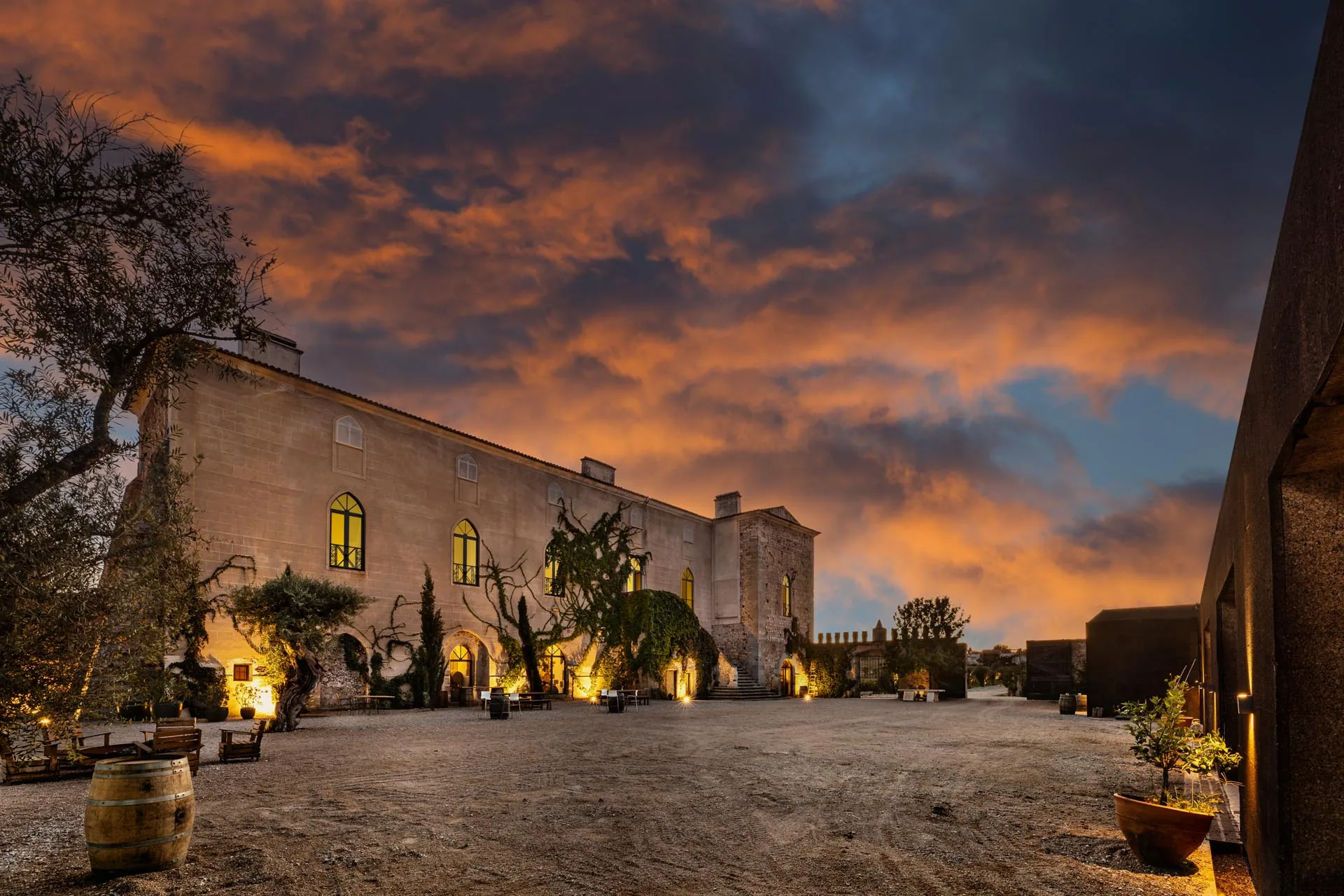 A stone manor with lit arched windows and a tower under an orange sunset sky, a dark modern building at the right