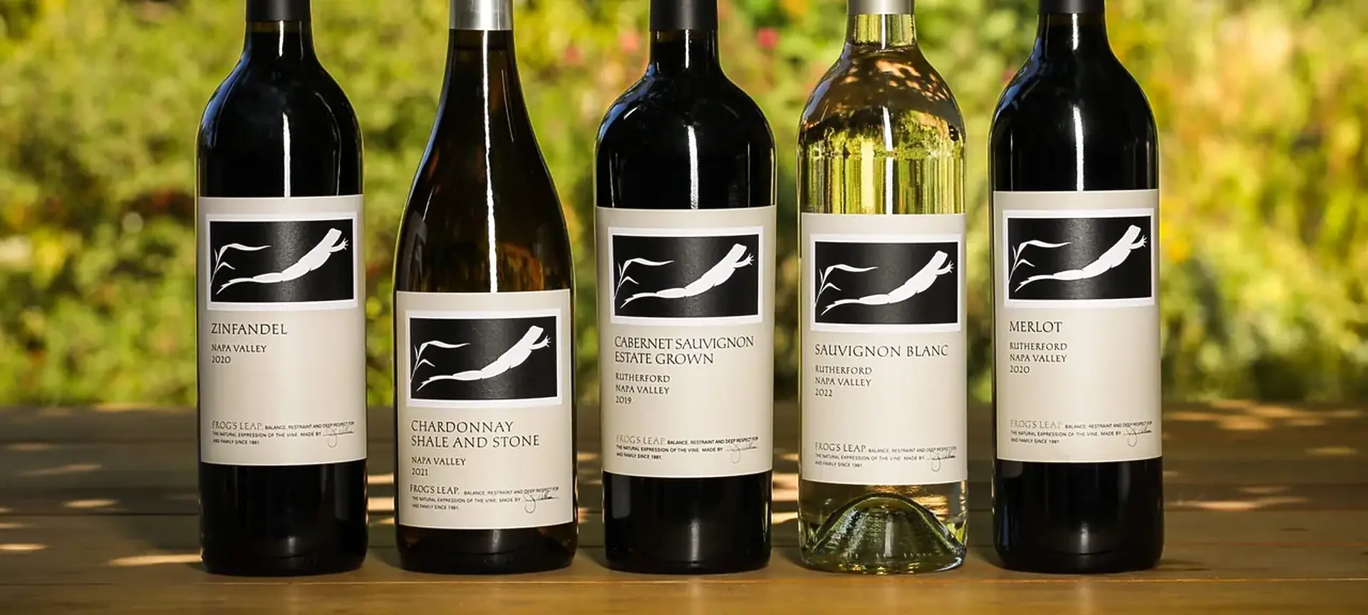 Five Frog's Leap bottles in a row on a wooden table: Zinfandel, Chardonnay, Cabernet Sauvignon, Sauvignon Blanc and Merlot