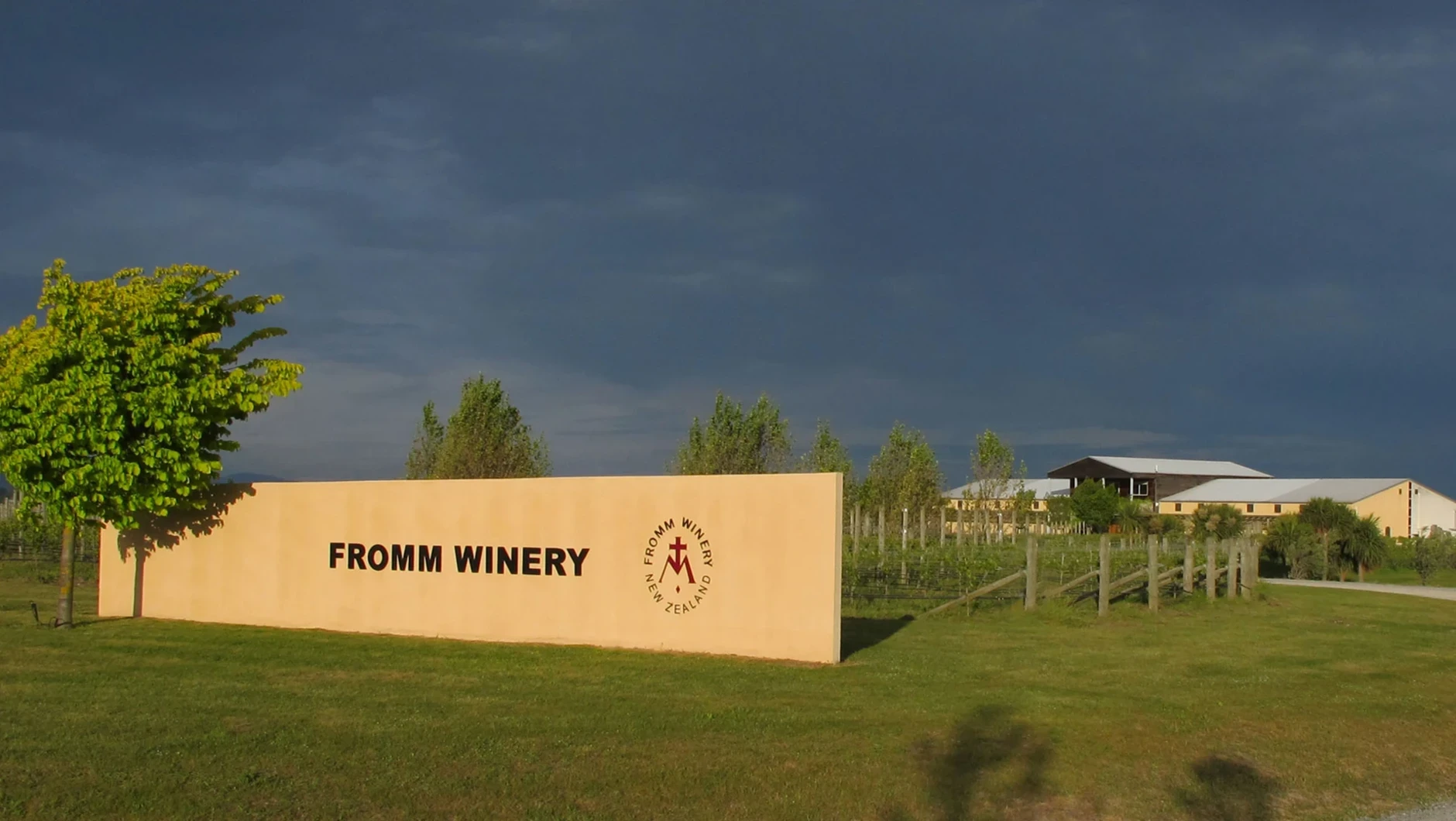 A tan wall sign reading Fromm Winery under a dark sky, with a green tree at left and winery buildings behind