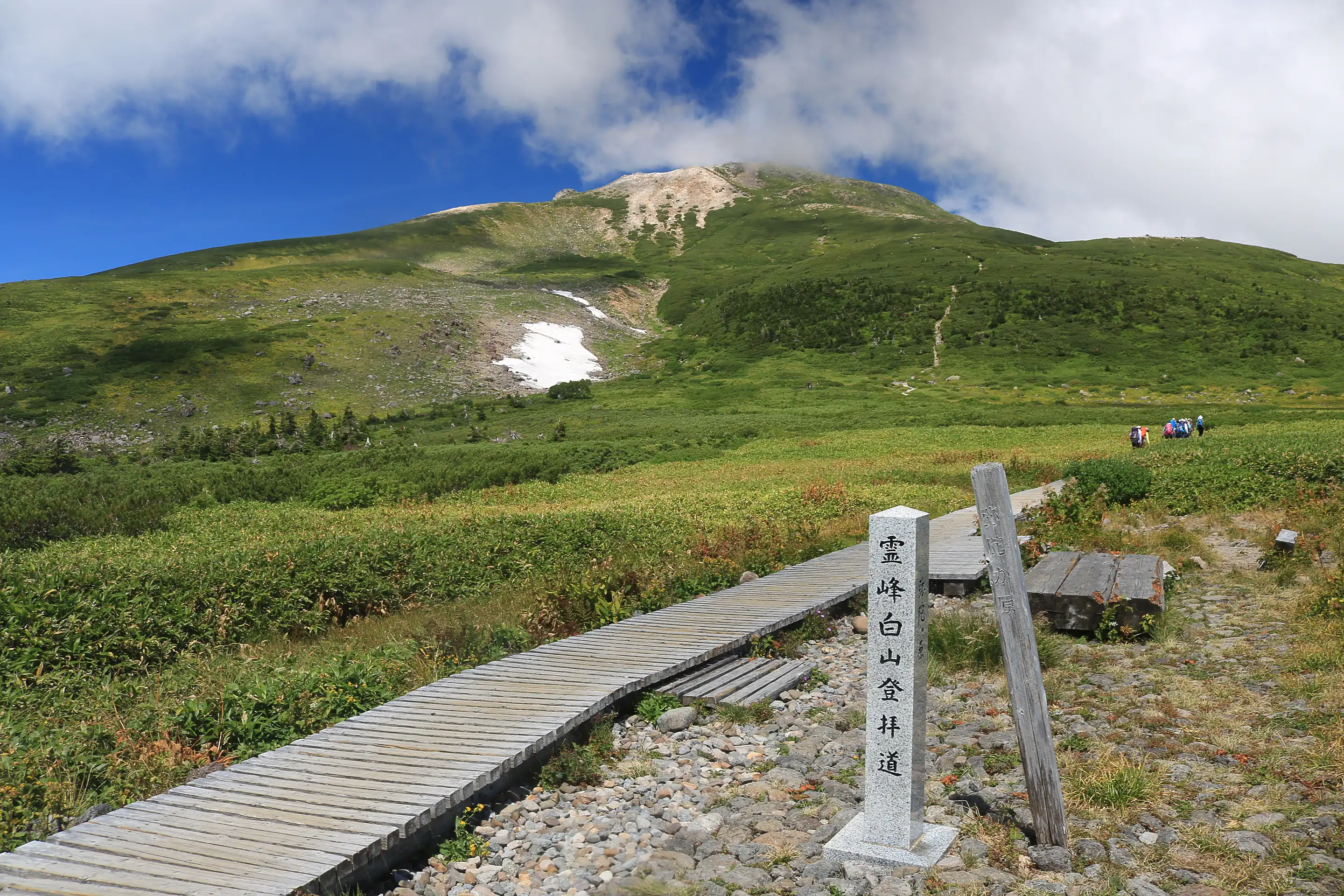 Mount Hakusan rising above grass and low shrubs, with a wooden boardwalk and a stone marker post at Midagahara in Ishikawa