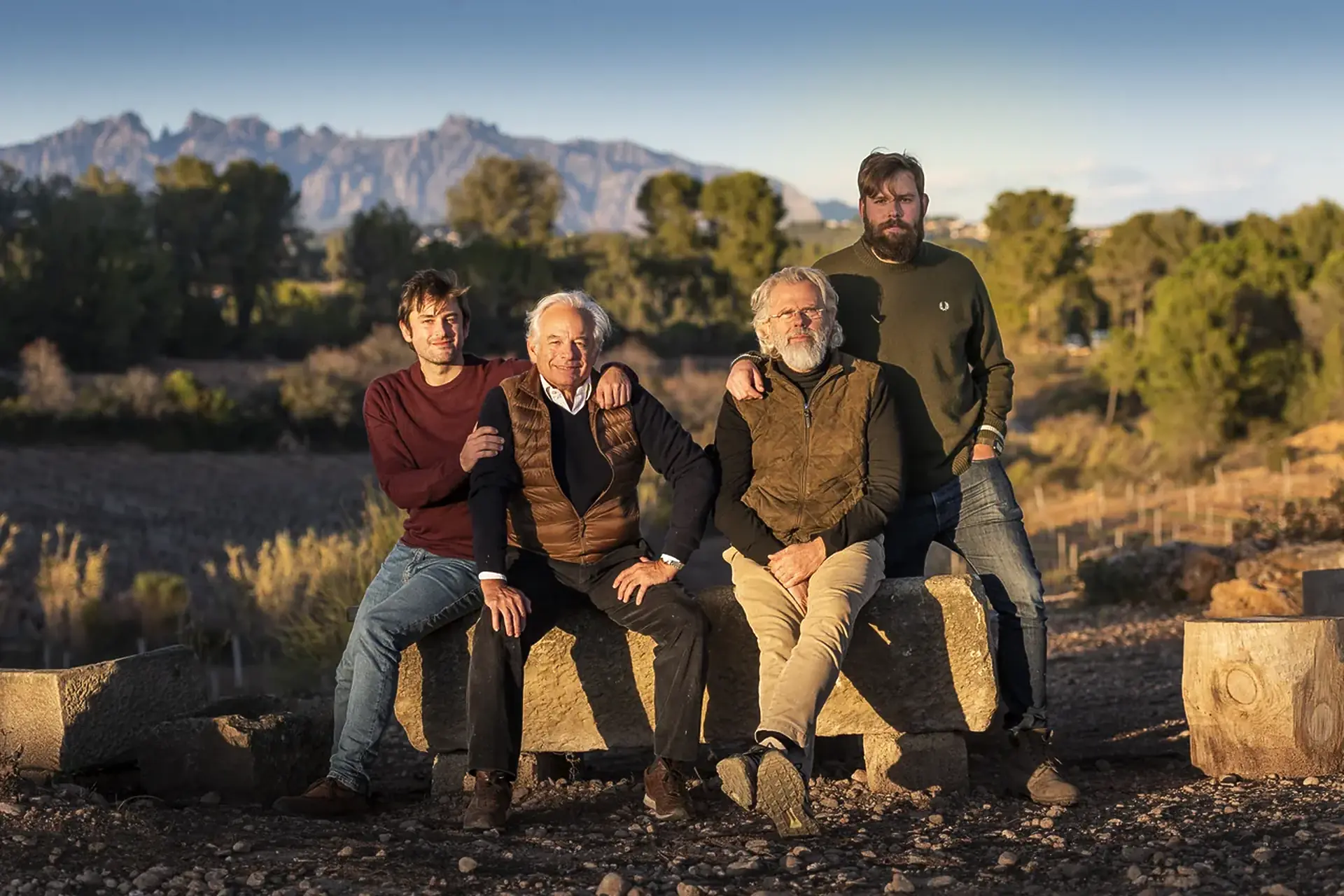 Four men sitting and standing on a stone block in dry scrubland, with a jagged mountain range behind in evening light