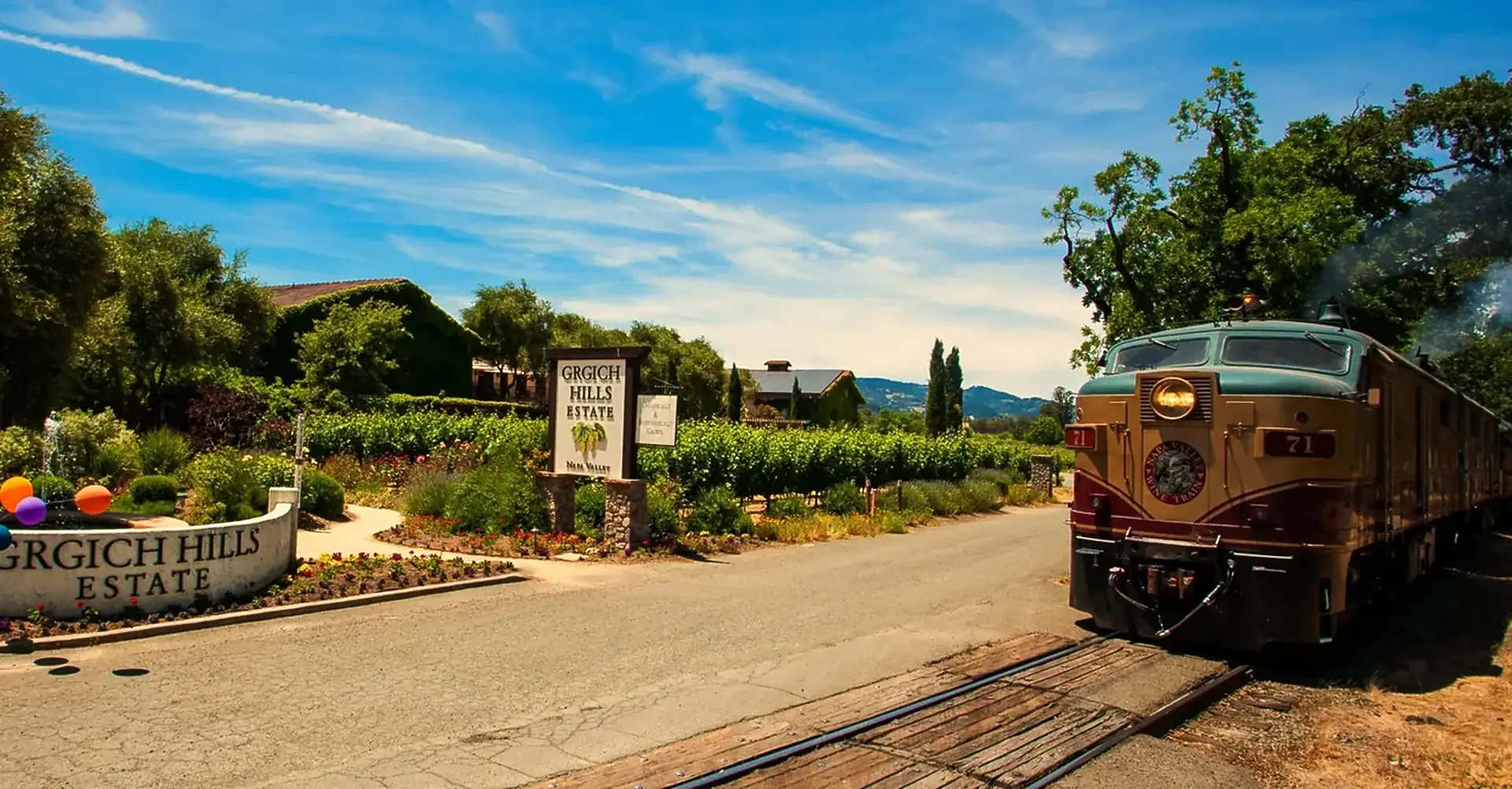A Grgich Hills Estate entrance sign beside a road, vines and flower beds behind, and a gold and green Napa Valley Wine Train locomotive at right