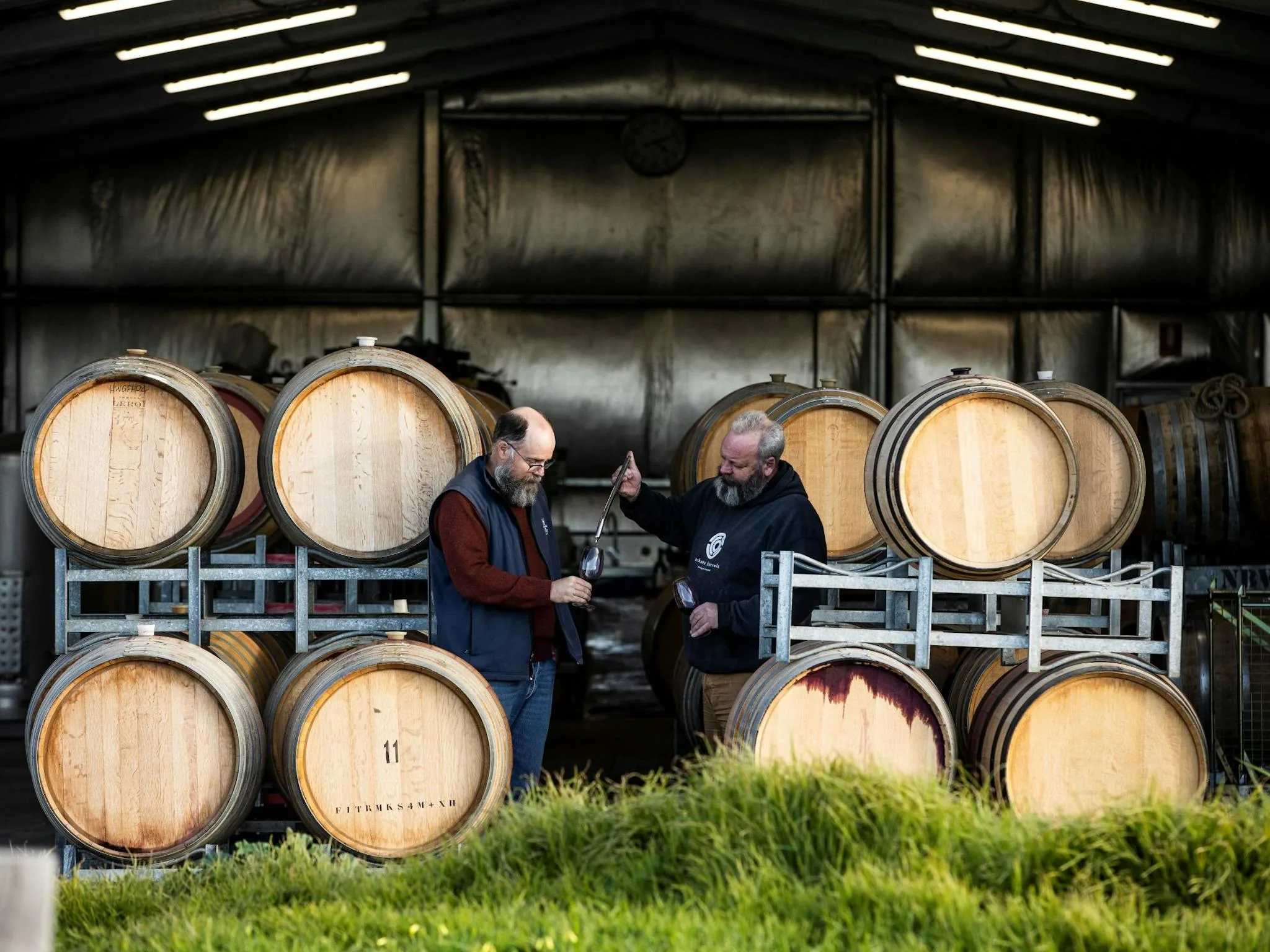Two bearded men tasting red wine in a shed, one holding a long sampling tube, among oak barrels stacked on galvanised racks
