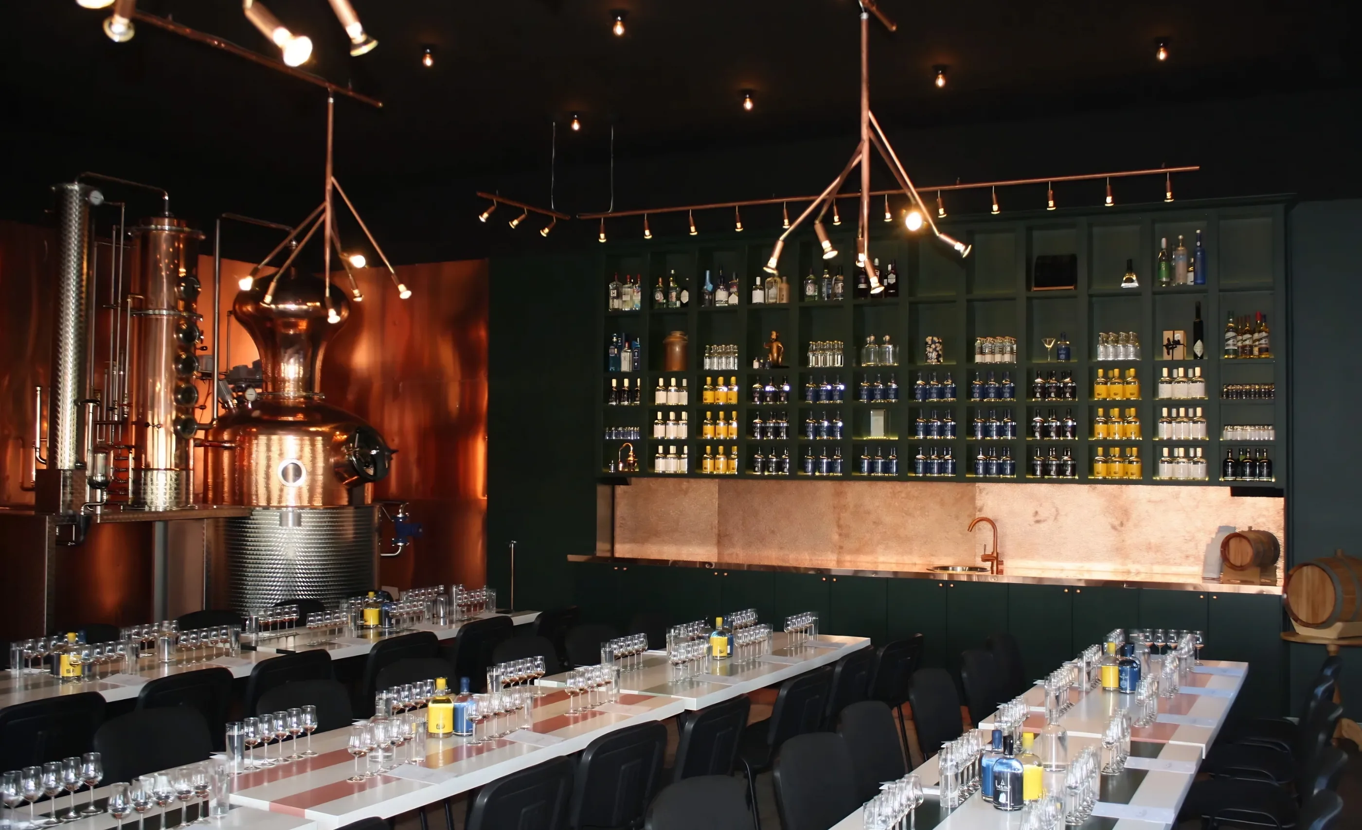 A tasting room with a copper pot still and column at left, a green shelf wall of bottles at the back, and long tables set with glasses