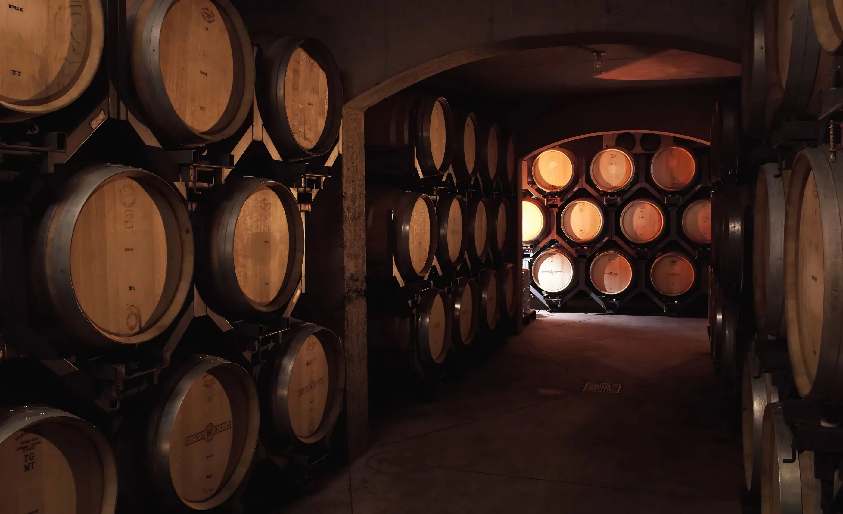 Oak barrels on steel racks lining a vaulted cellar passage, the barrel heads marked Hidden Bench, with a lit room beyond