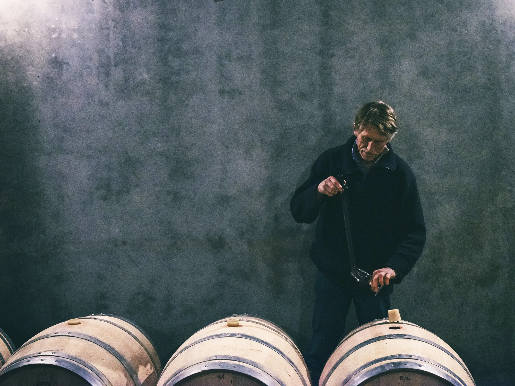 A winemaker in a dark jacket drawing wine from a barrel with a long sampling tube, against a bare concrete wall