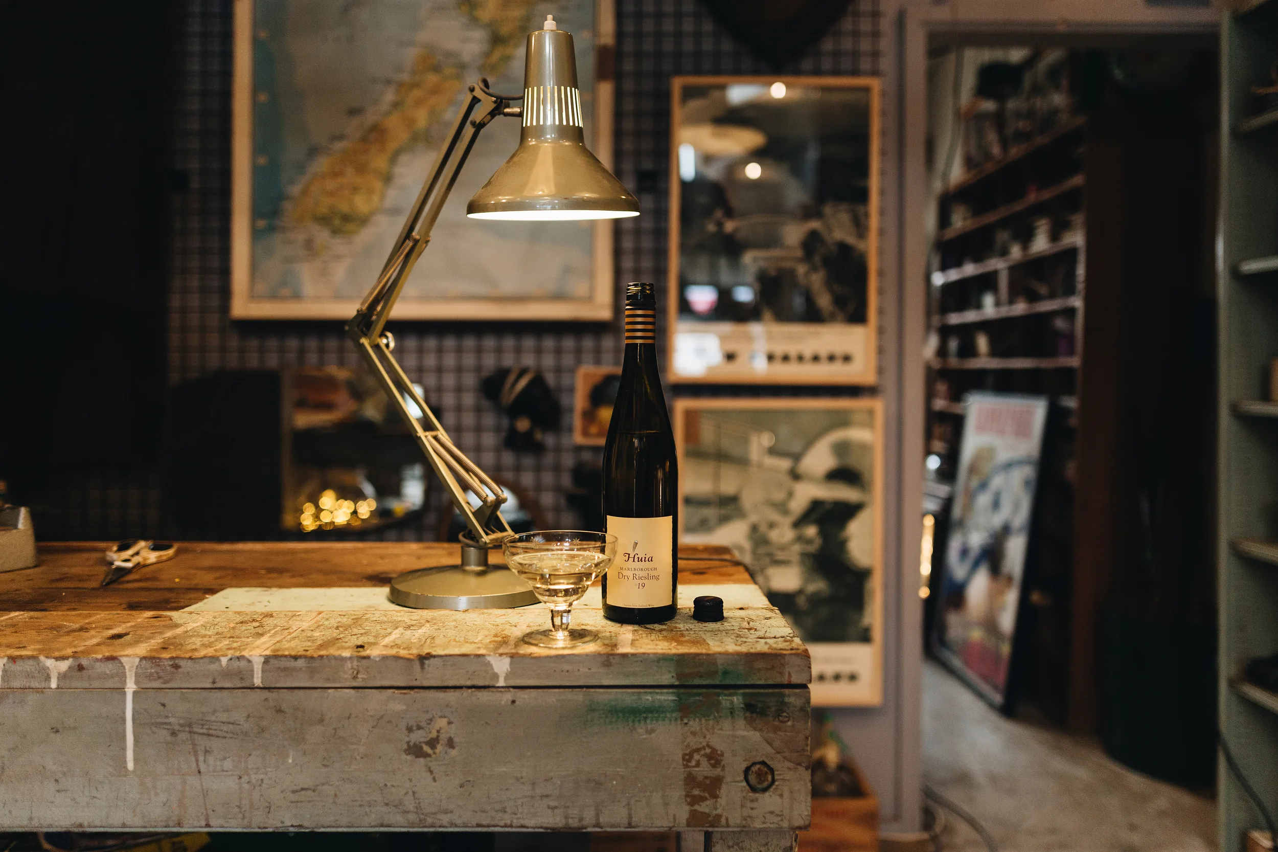 A bottle of Huia Marlborough Dry Riesling and a coupe glass on a worn workbench under an anglepoise lamp, a map of New Zealand behind
