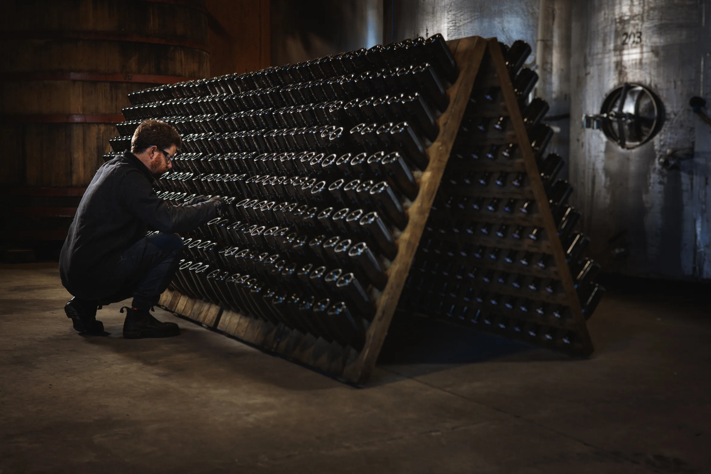 A man crouching to hand-riddle bottles on a large A-frame rack of sparkling wine in the Huia cellar