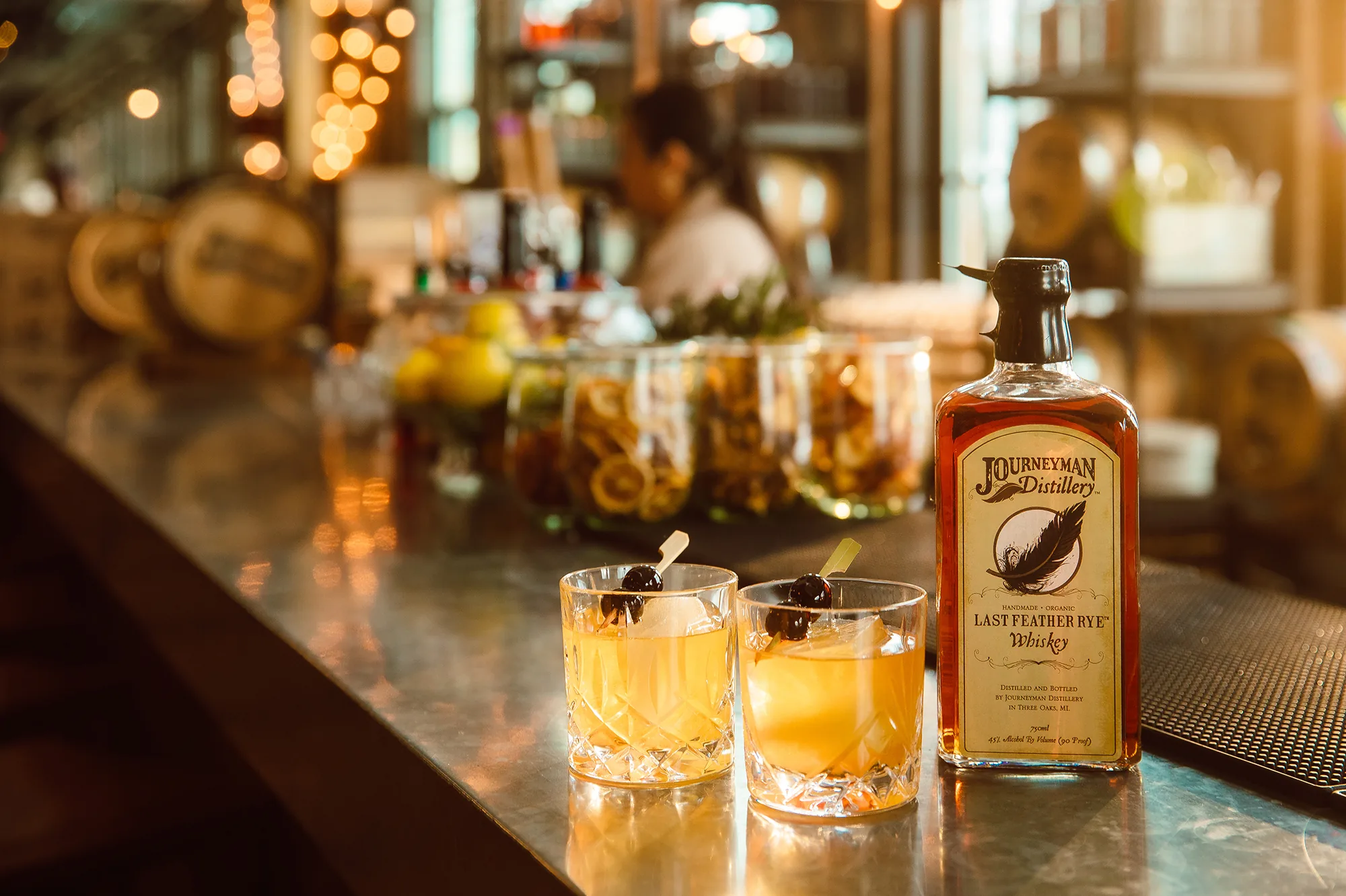 A bottle labelled Journeyman Distillery Last Feather Rye Whiskey beside two cherry-garnished drinks on a metal bar counter
