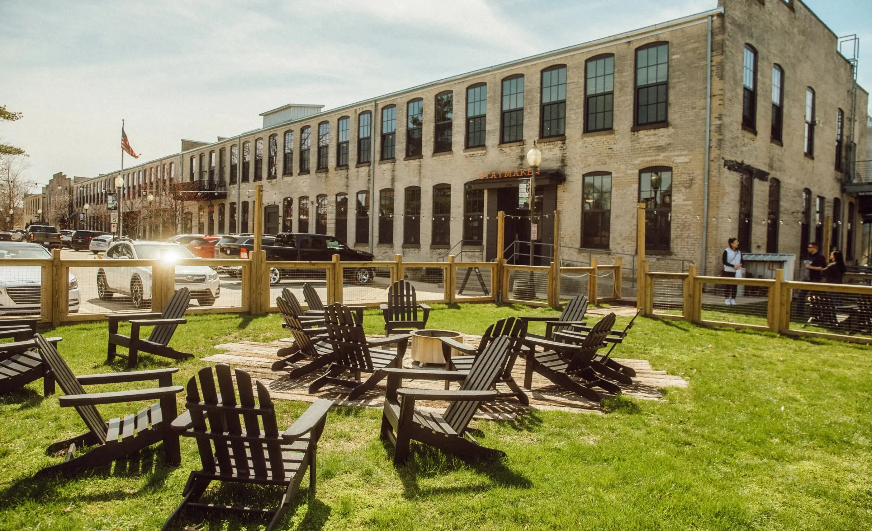Black Adirondack chairs around a fire pit on a lawn in front of a long brick building with arched windows