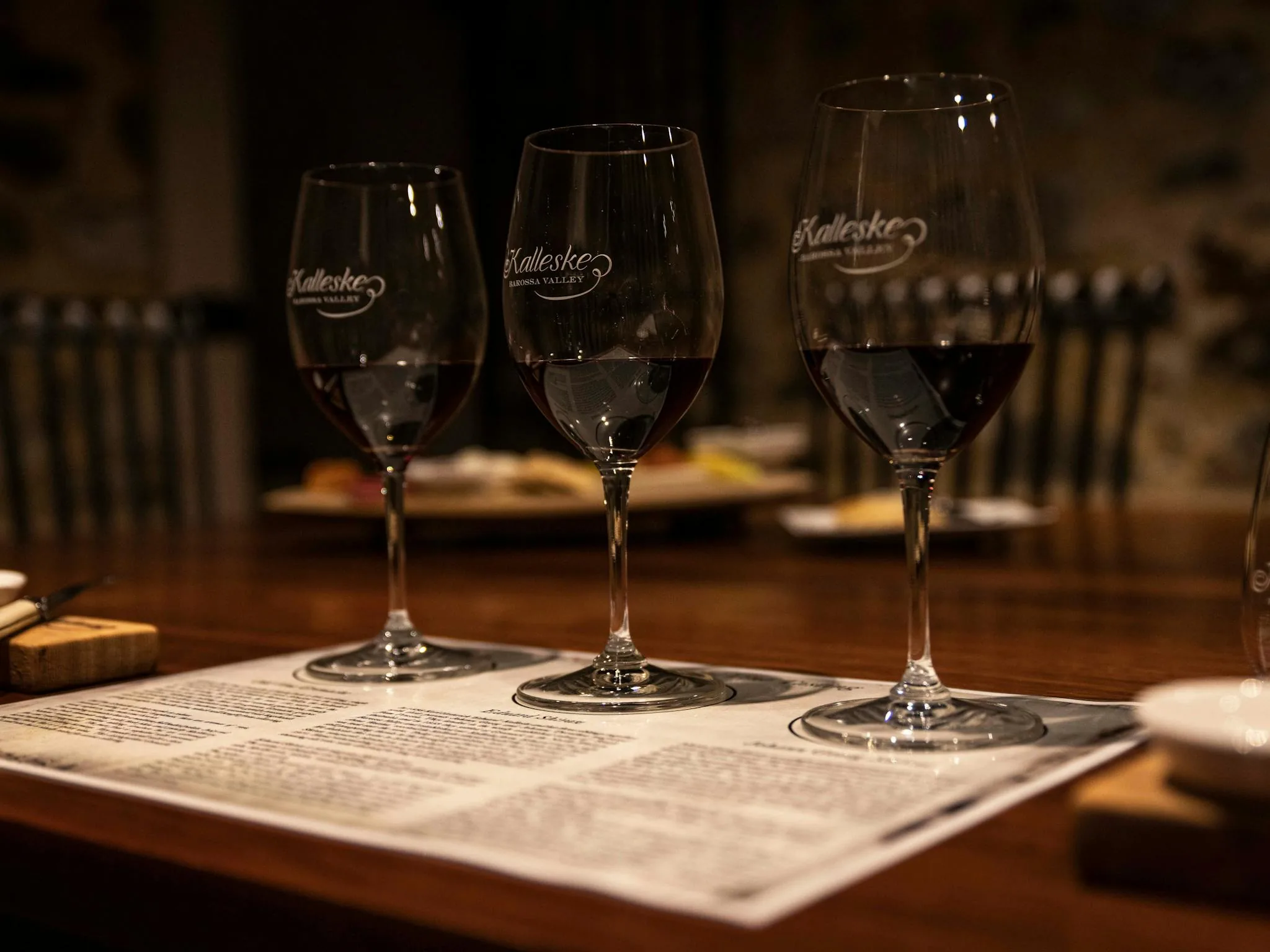 Three wine glasses etched with the Kalleske Barossa Valley logo, part-filled with red wine, on a printed tasting sheet