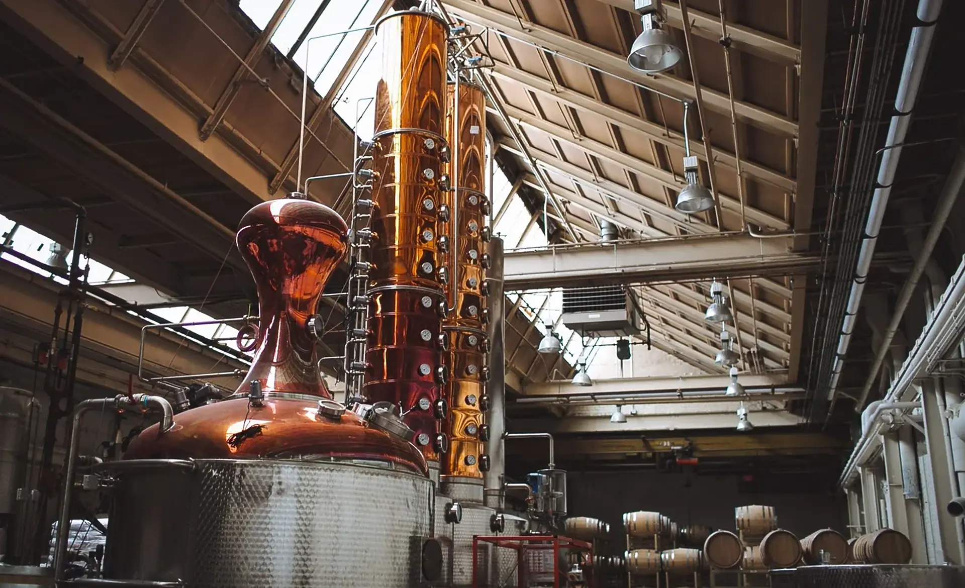 A copper pot still and column still under a high skylit roof, with barrels racked against the far wall