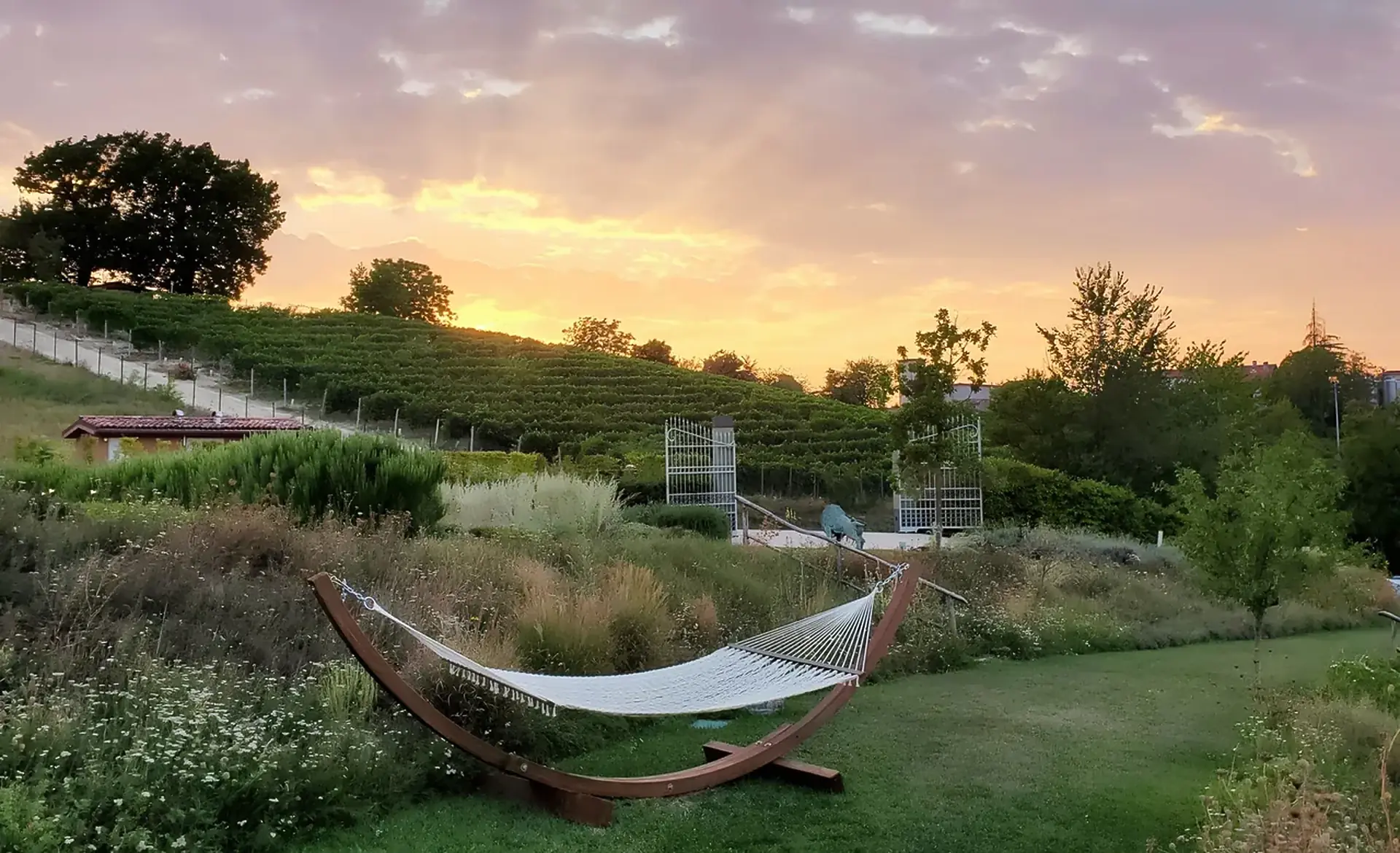 A hammock on a curved wooden stand in a garden of grasses and wildflowers, with a vine-covered hill under a pink sunset sky