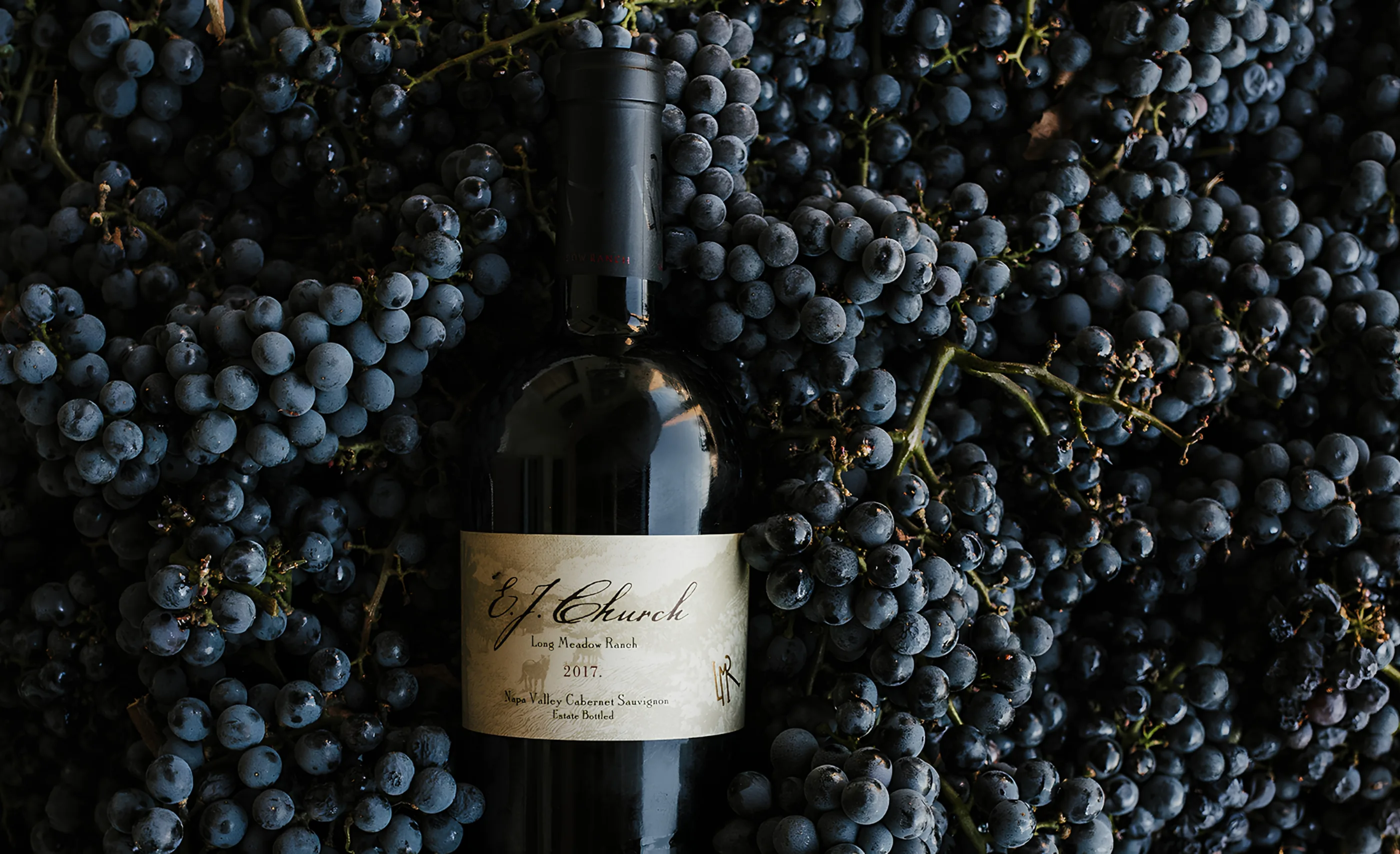 A bottle labelled E. J. Church Long Meadow Ranch Napa Valley Cabernet Sauvignon 2017 lying on heaped dark grape clusters
