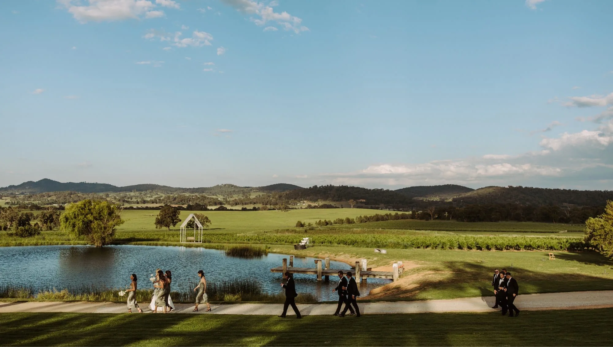 Wedding guests in suits and green dresses walking along a gravel track beside a dam, with vines, paddocks and low hills beyond