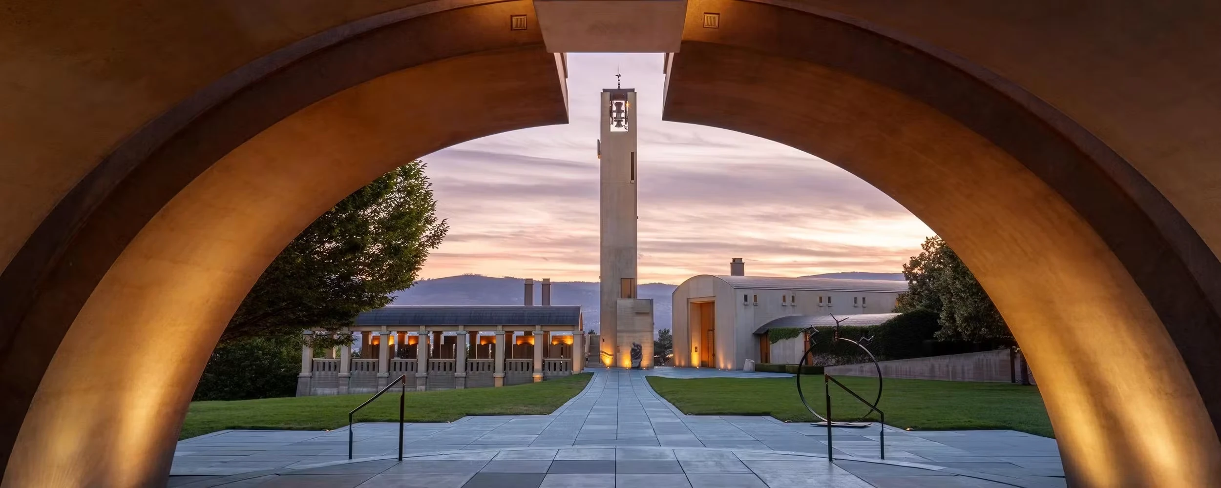 Looking through a large curved stone arch at a bell tower, a colonnaded pavilion and a lawn at dusk, with hills behind