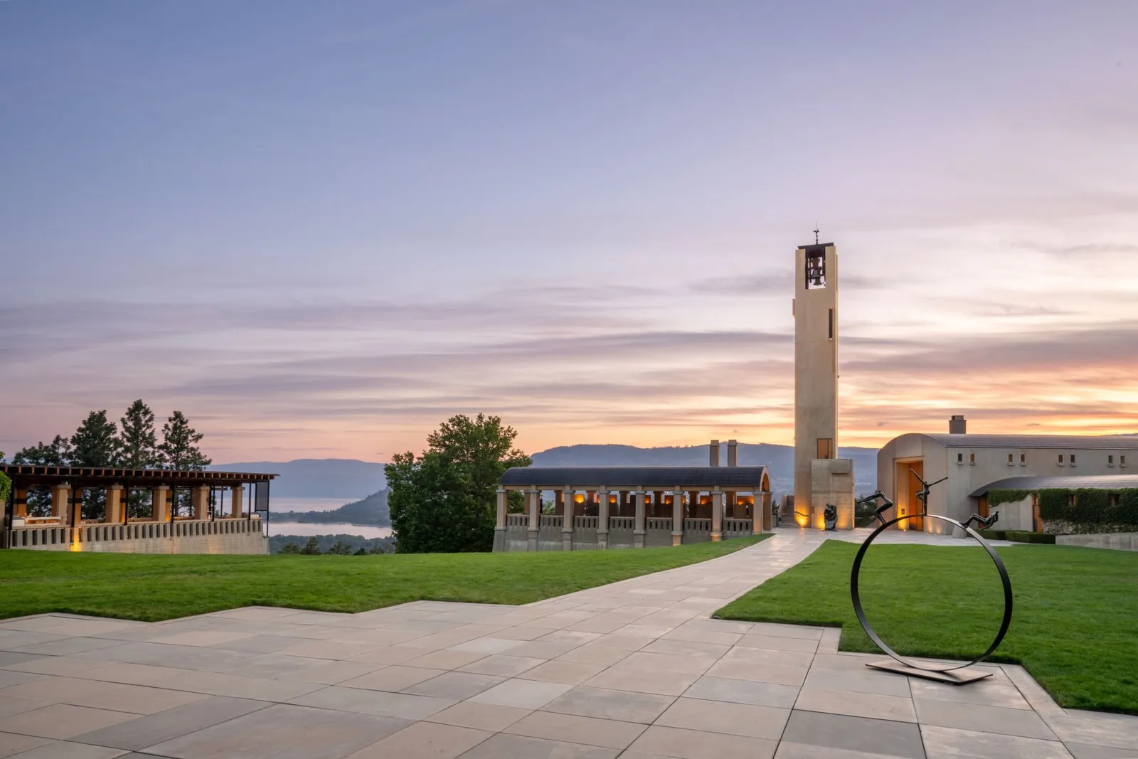 A stone bell tower, a colonnaded pavilion and a ring-shaped metal sculpture on a lawn at dusk, hills and water beyond