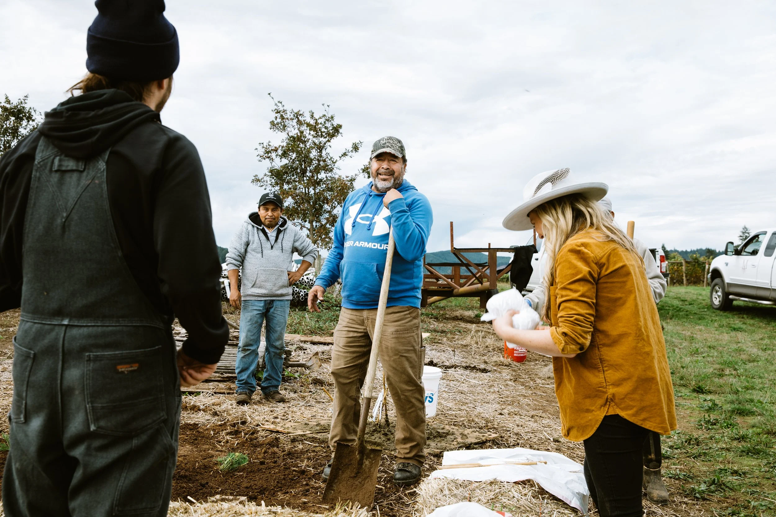 Five people gathered on bare ground at a planting or compost session, one leaning on a shovel, a white pickup truck behind