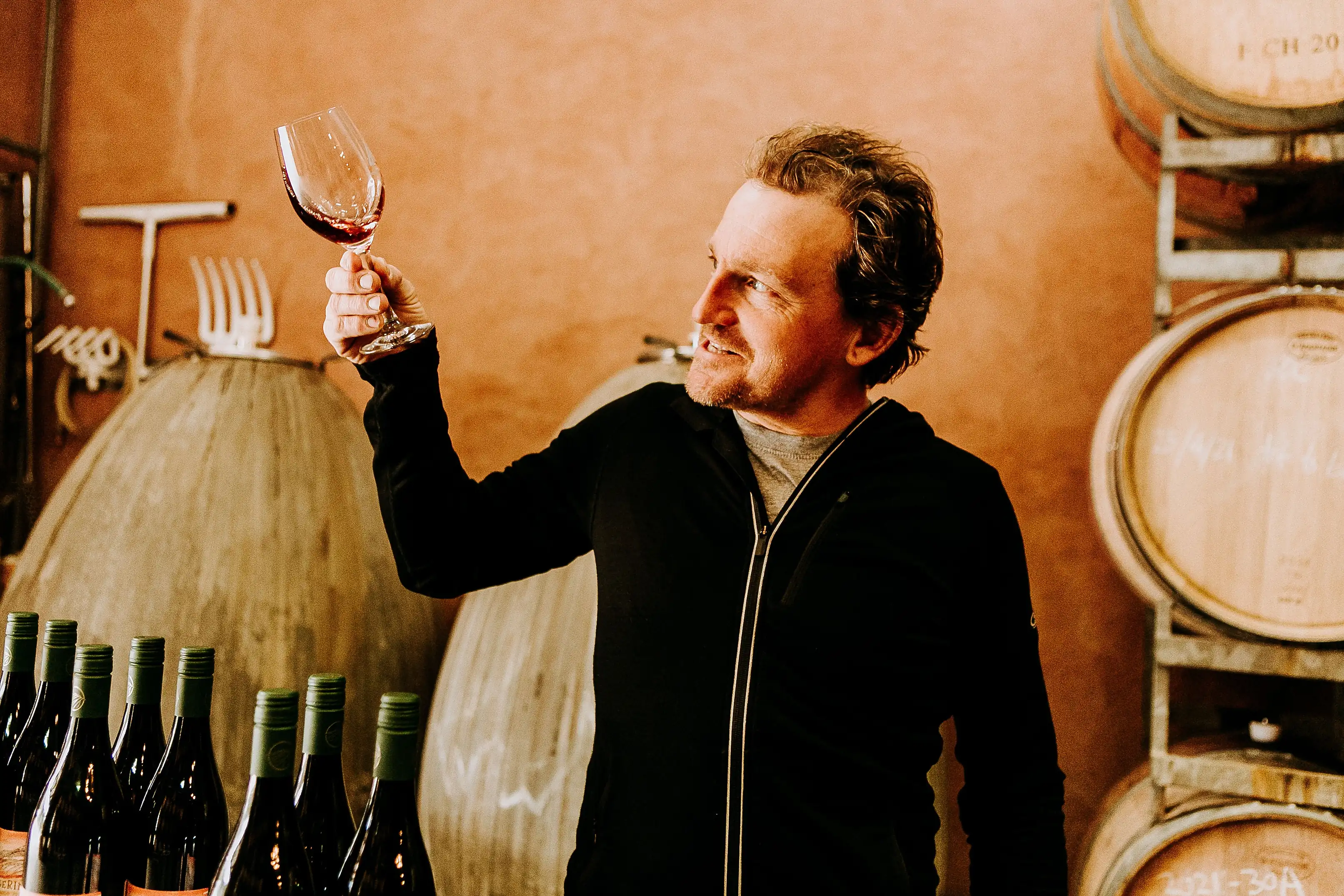 A winemaker holding a glass of red wine up to the light in the cellar at Ngeringa, concrete eggs and oak barrels behind