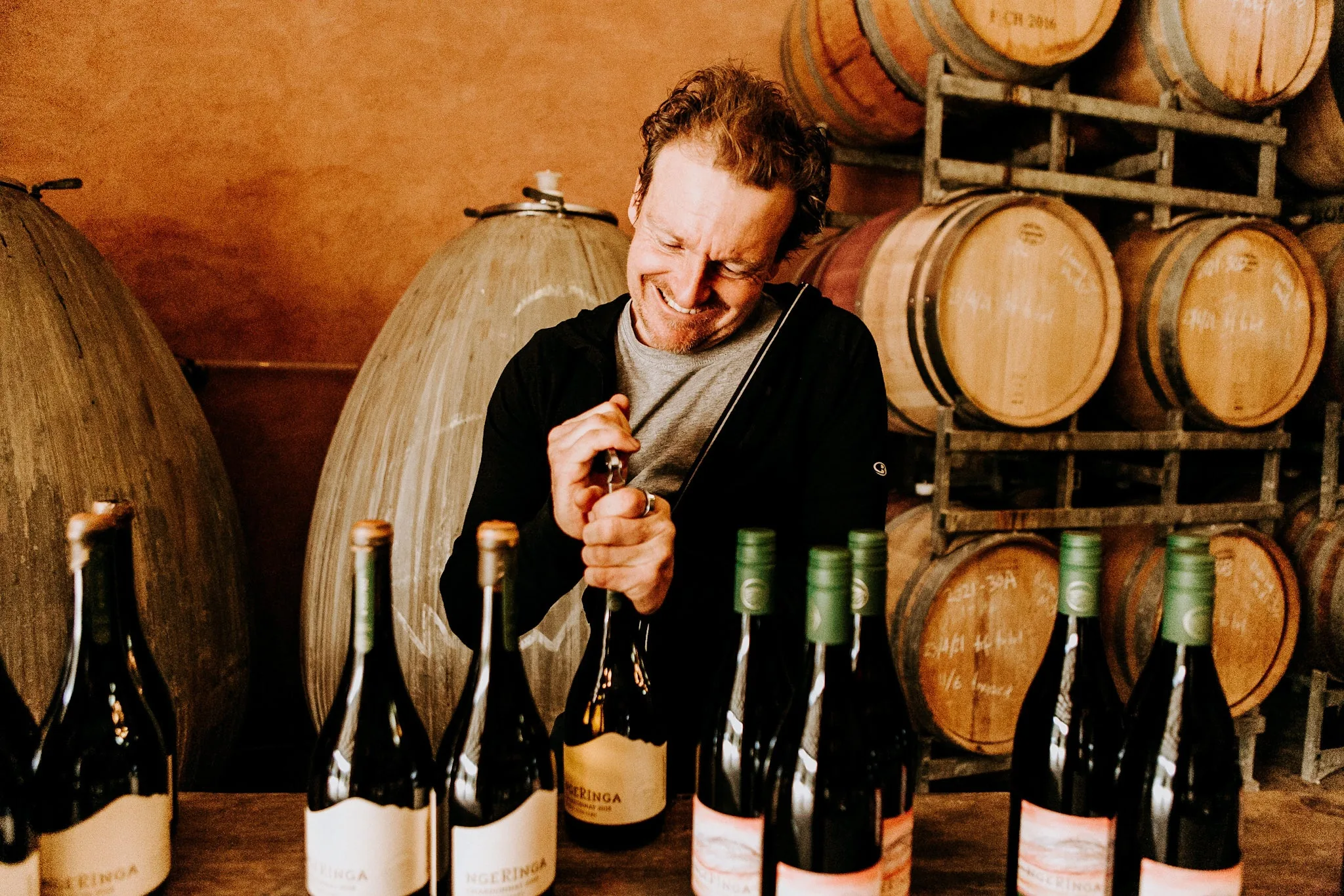 A winemaker opening a bottle with a corkscrew among rows of Ngeringa bottles, concrete eggs and oak barrels behind
