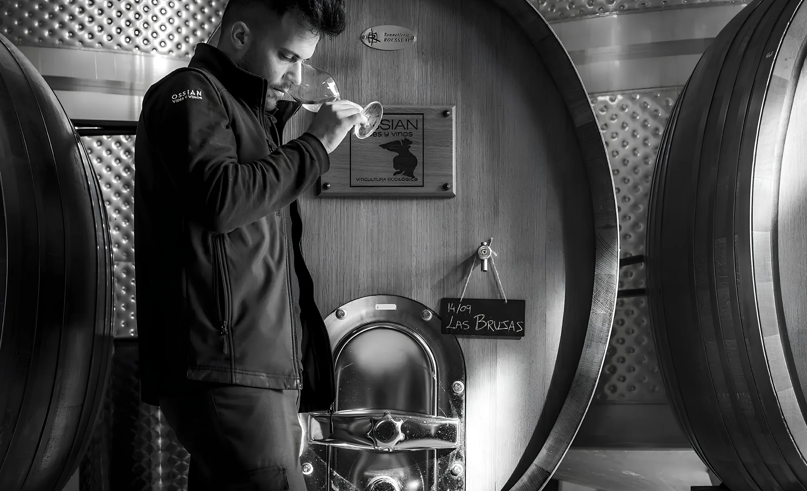 A man in an Ossian jacket nosing a glass of wine beside a large oak vat with an Ossian plaque, in black and white