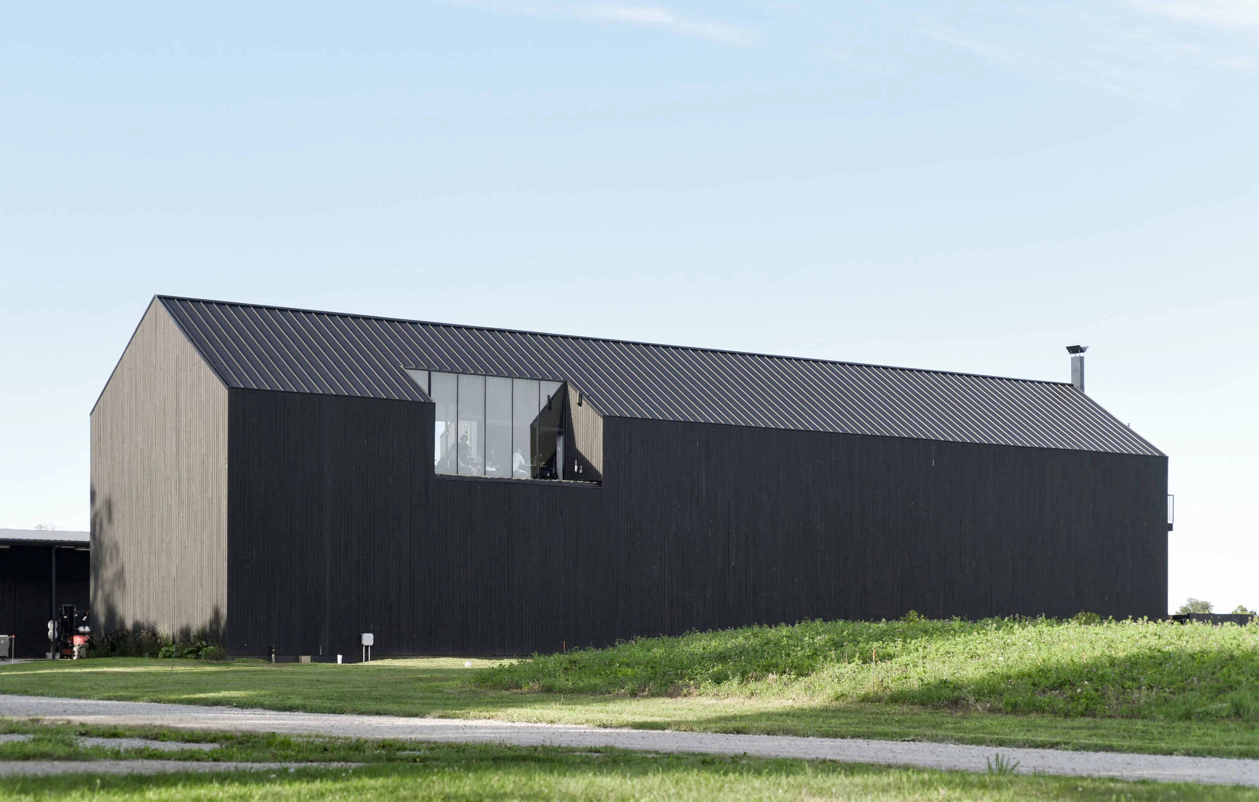 A black timber-clad barn-shaped building with a dark metal roof and a tall window, behind a grassy bank, at Pearl Morissette