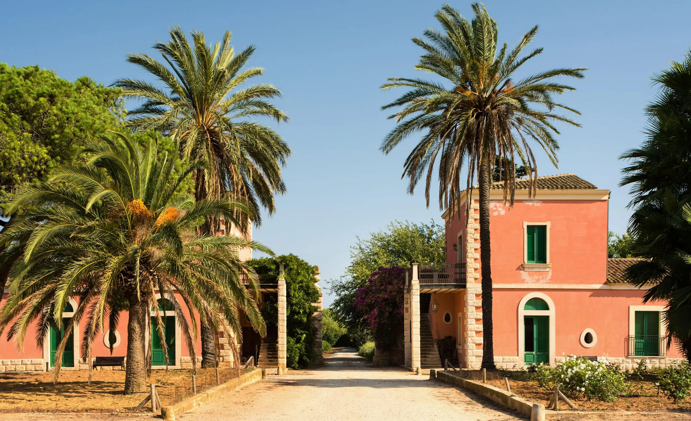 A coral-pink building with green shutters and arched windows, flanked by tall palm trees along a gravel drive, under a clear sky