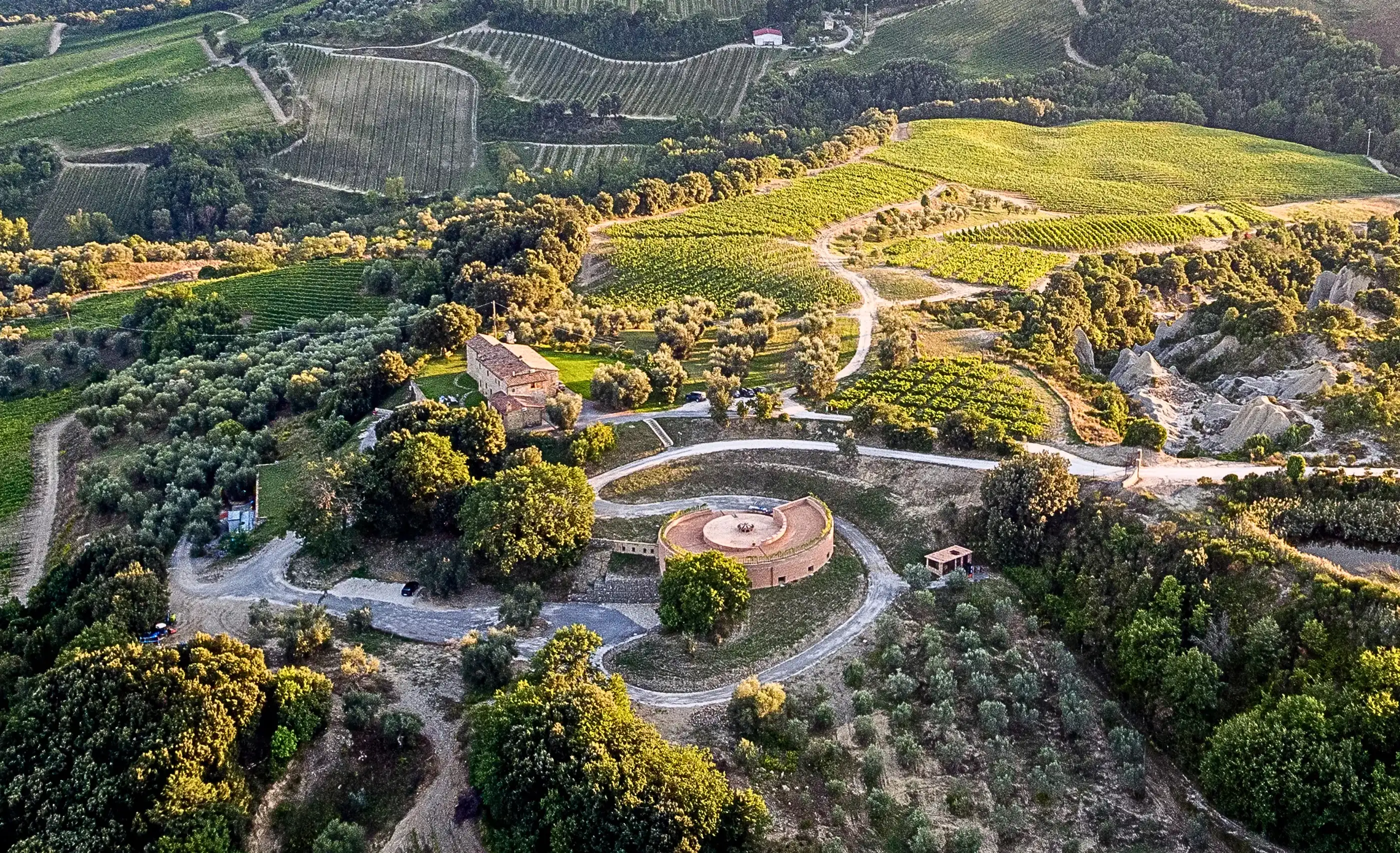 Aerial view of a round building among olive groves, vineyards and eroded clay slopes, at Podere Le Ripi