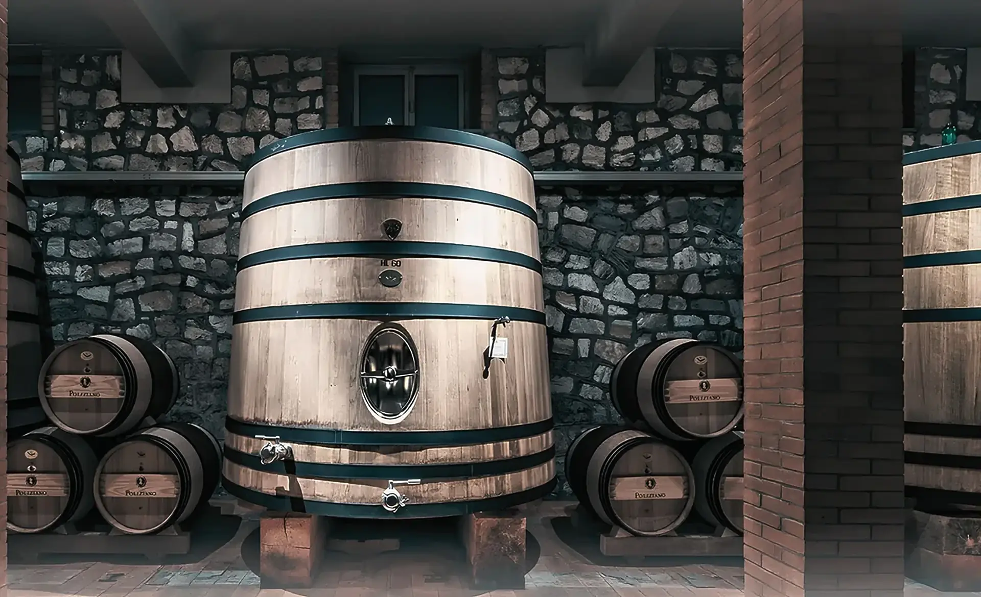 A large conical oak cask between stacks of small barrels marked Poliziano, in a stone-walled cellar with a brick pillar