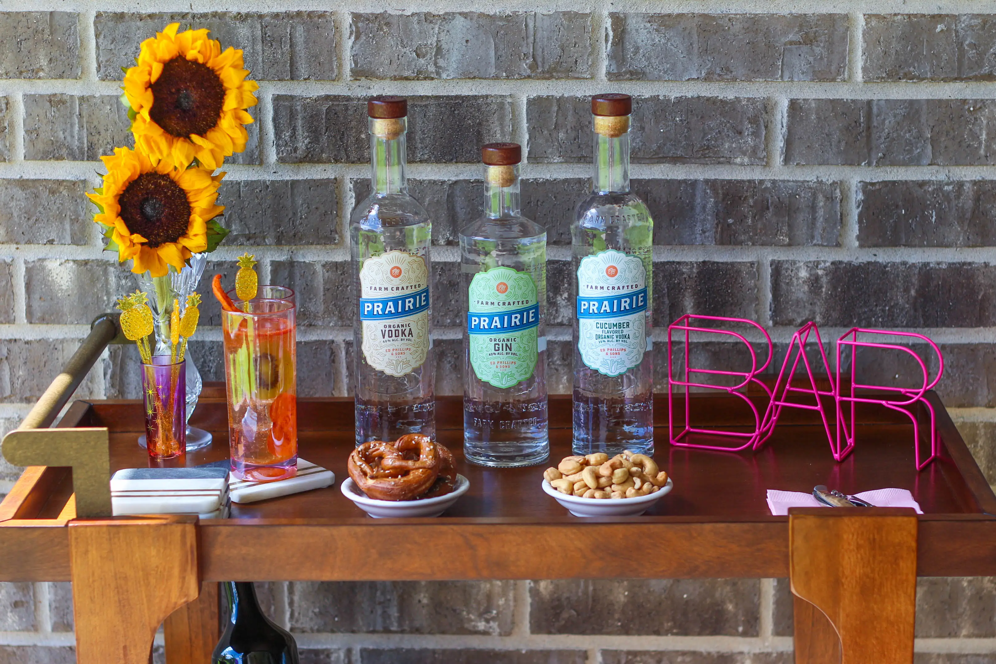 Three Prairie bottles, vodka, gin and cucumber vodka, on a wooden bar cart with sunflowers, snacks and a pink BAR sign