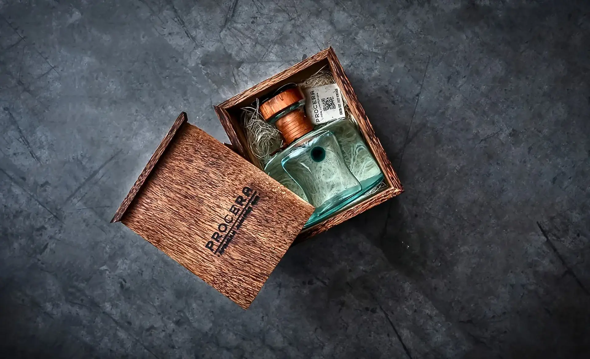 A Procera bottle in an open wooden box with the PROCERA name burned into the lid, on a dark slate surface