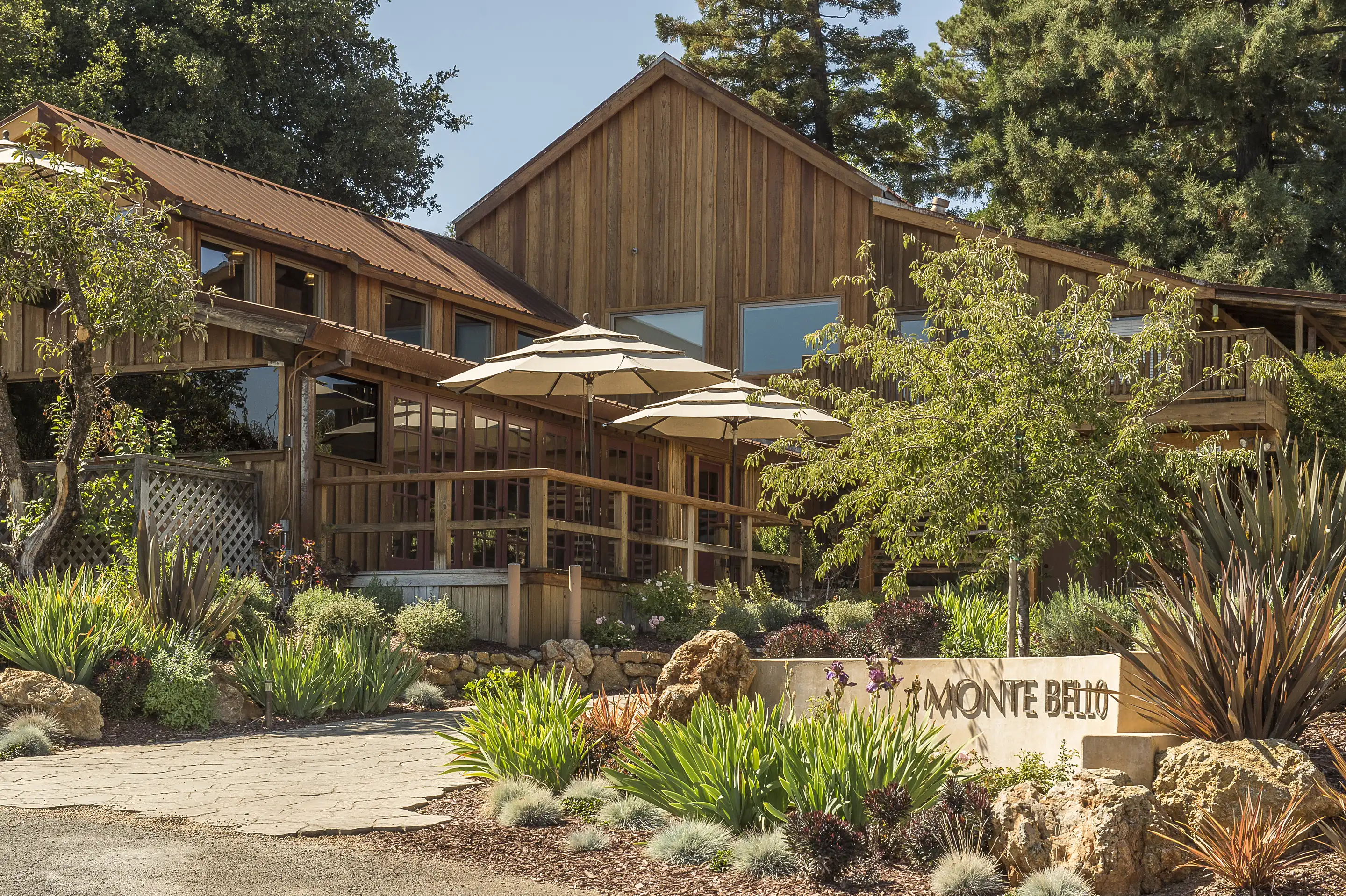 Timber tasting room with umbrellas on its terrace and a Monte Bello sign among rock and grass plantings, at Ridge Vineyards