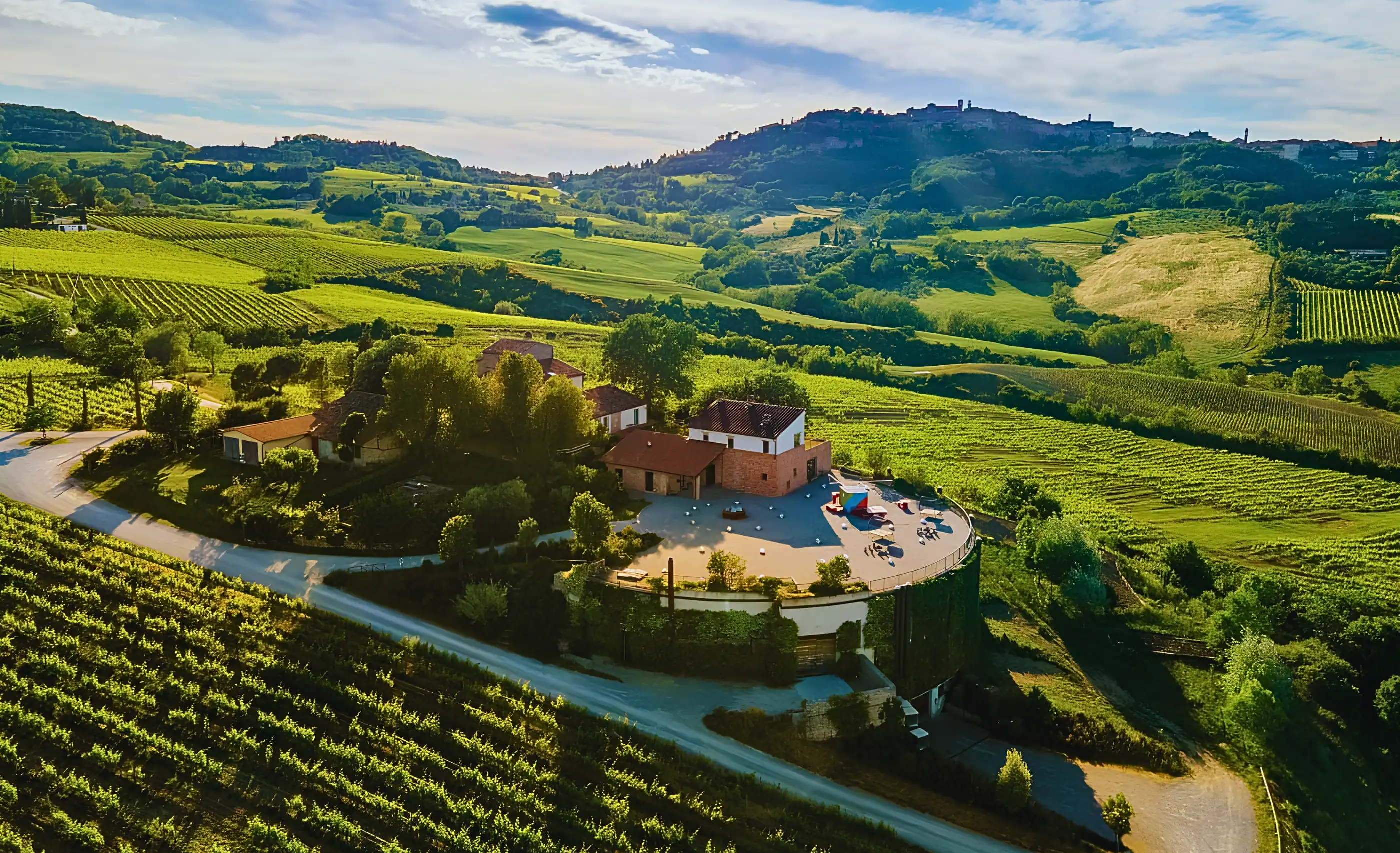Aerial view of a hilltop winery with a round terrace among green vineyards, with a hill town on the ridge behind