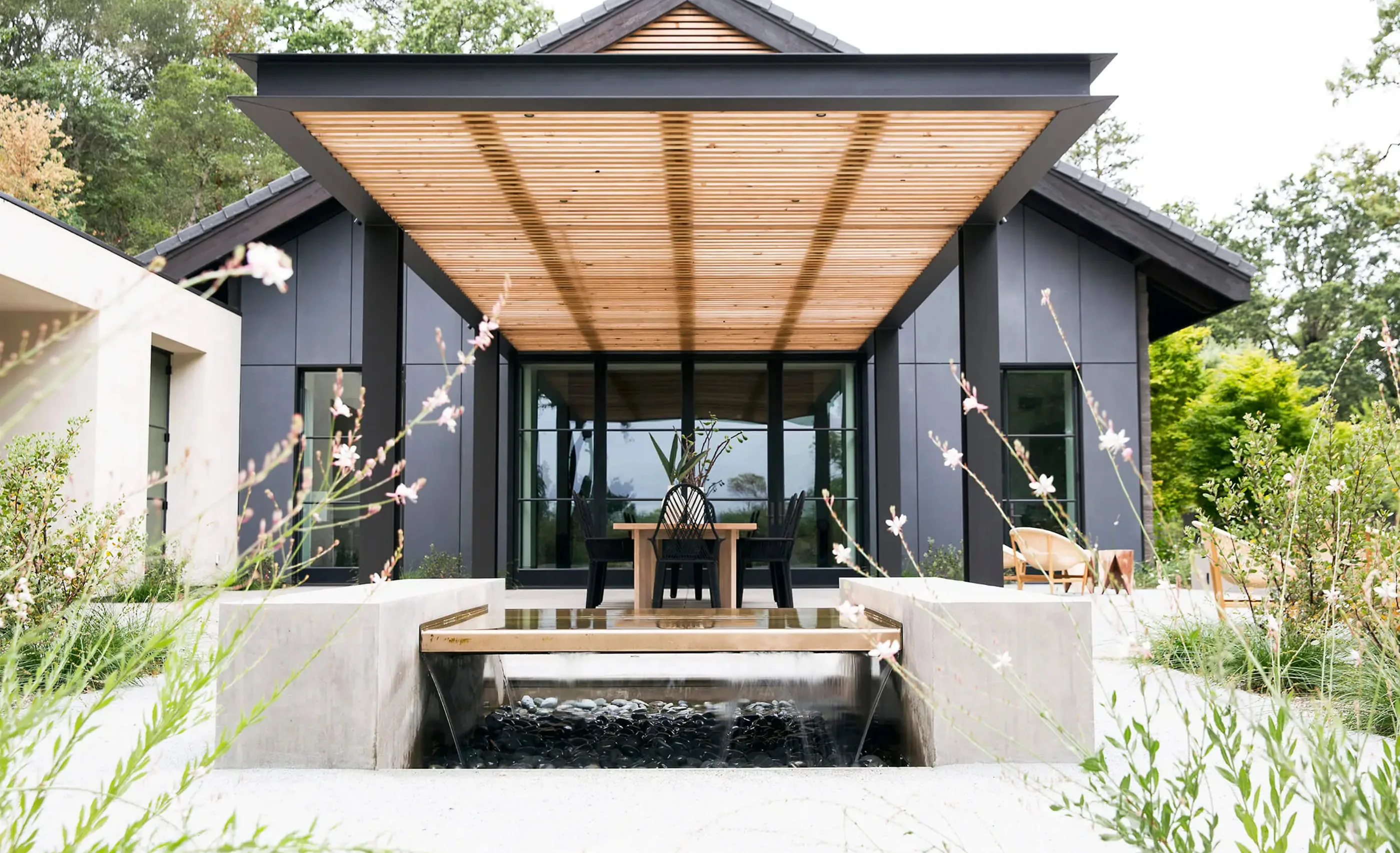 Dark-clad building with a slatted timber canopy, a water channel over black pebbles and flowering grasses, at Sinegal Estate