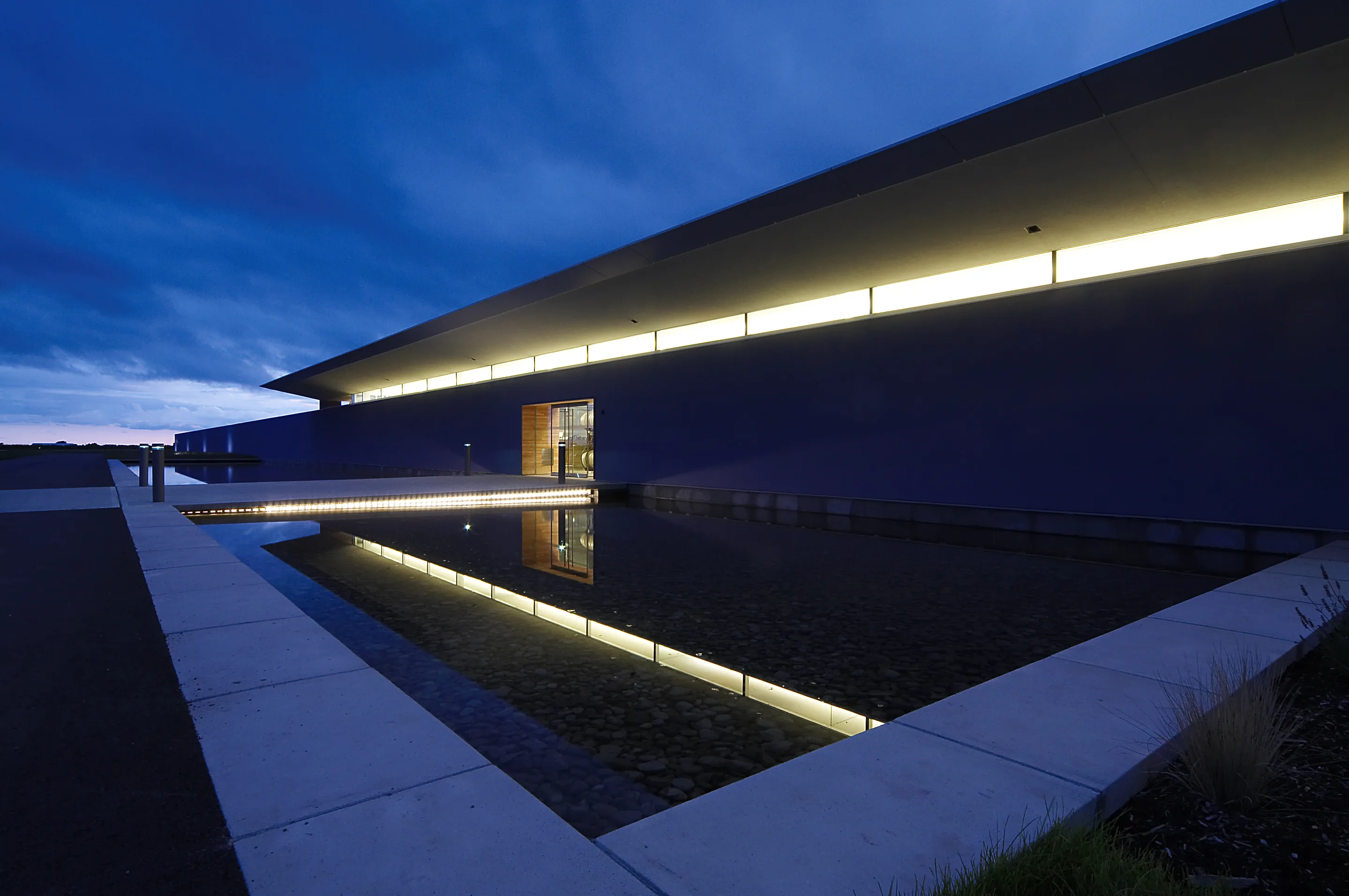 Long, low modern building lit from within at dusk, with a still rectangular pool of dark water in the foreground
