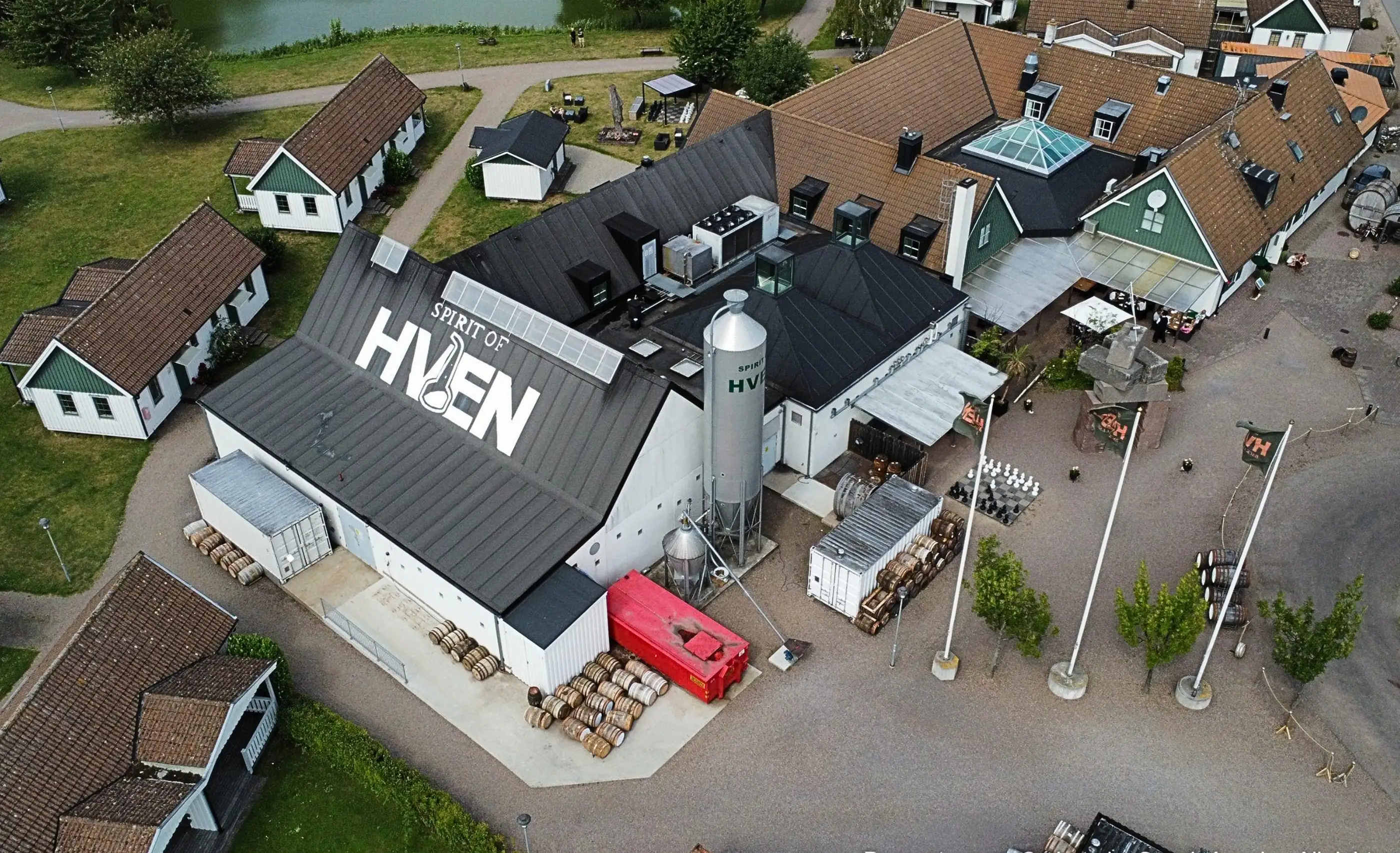 Aerial view of a dark roof lettered Spirit of Hven, with a silo, stacked barrels, a pond and white cottages with brown tiled roofs