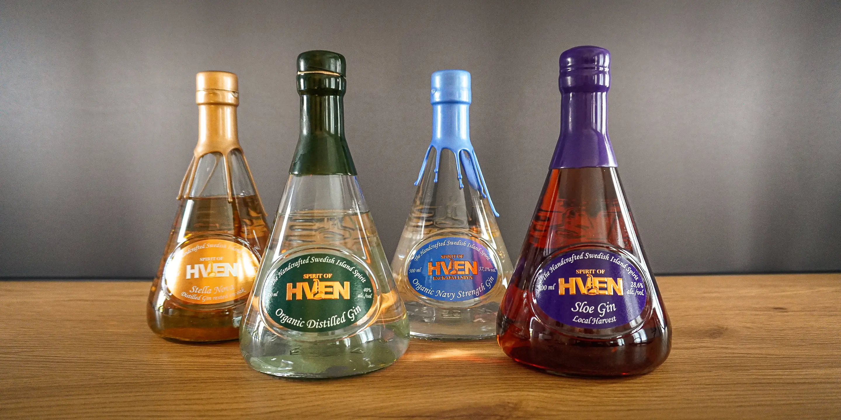 Four conical Spirit of Hven bottles on a wooden surface, with gold, green, blue and purple closures