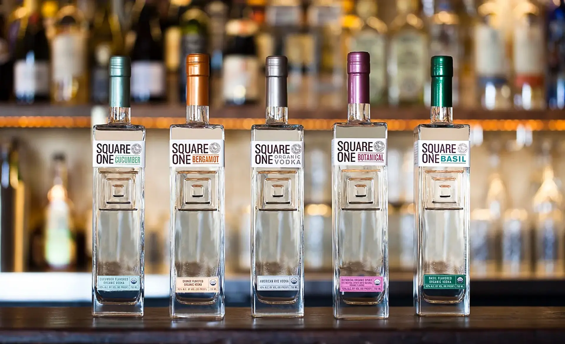 Five square glass bottles labelled Square One, with Cucumber, Bergamot, Botanical and Basil among them, lined up on a bar
