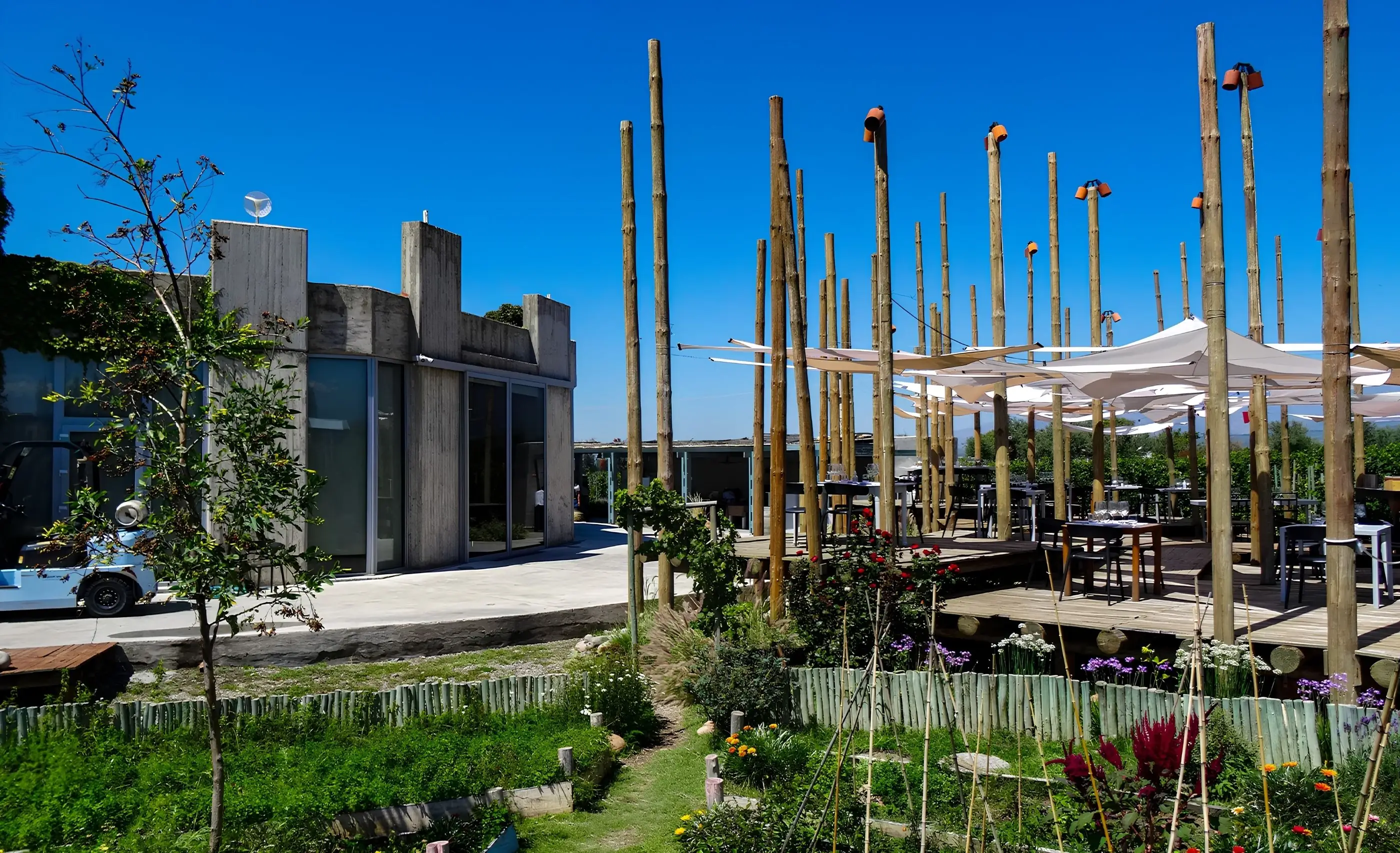 A concrete building beside a timber dining deck under white canopies and tall bamboo poles, with a flower garden in front