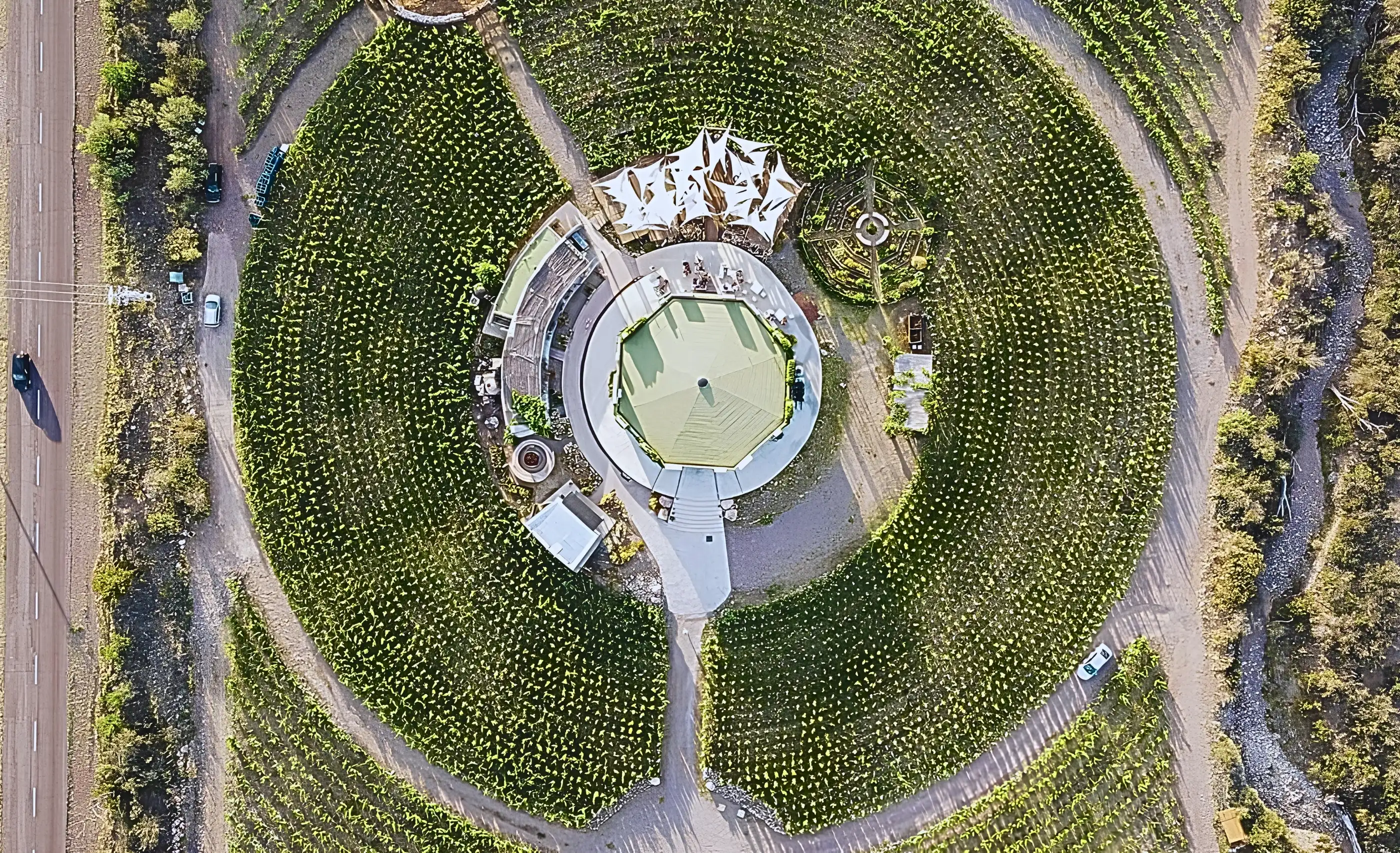Overhead view of circular vineyard rows around a round green-roofed building, a road at left and a stony channel at right
