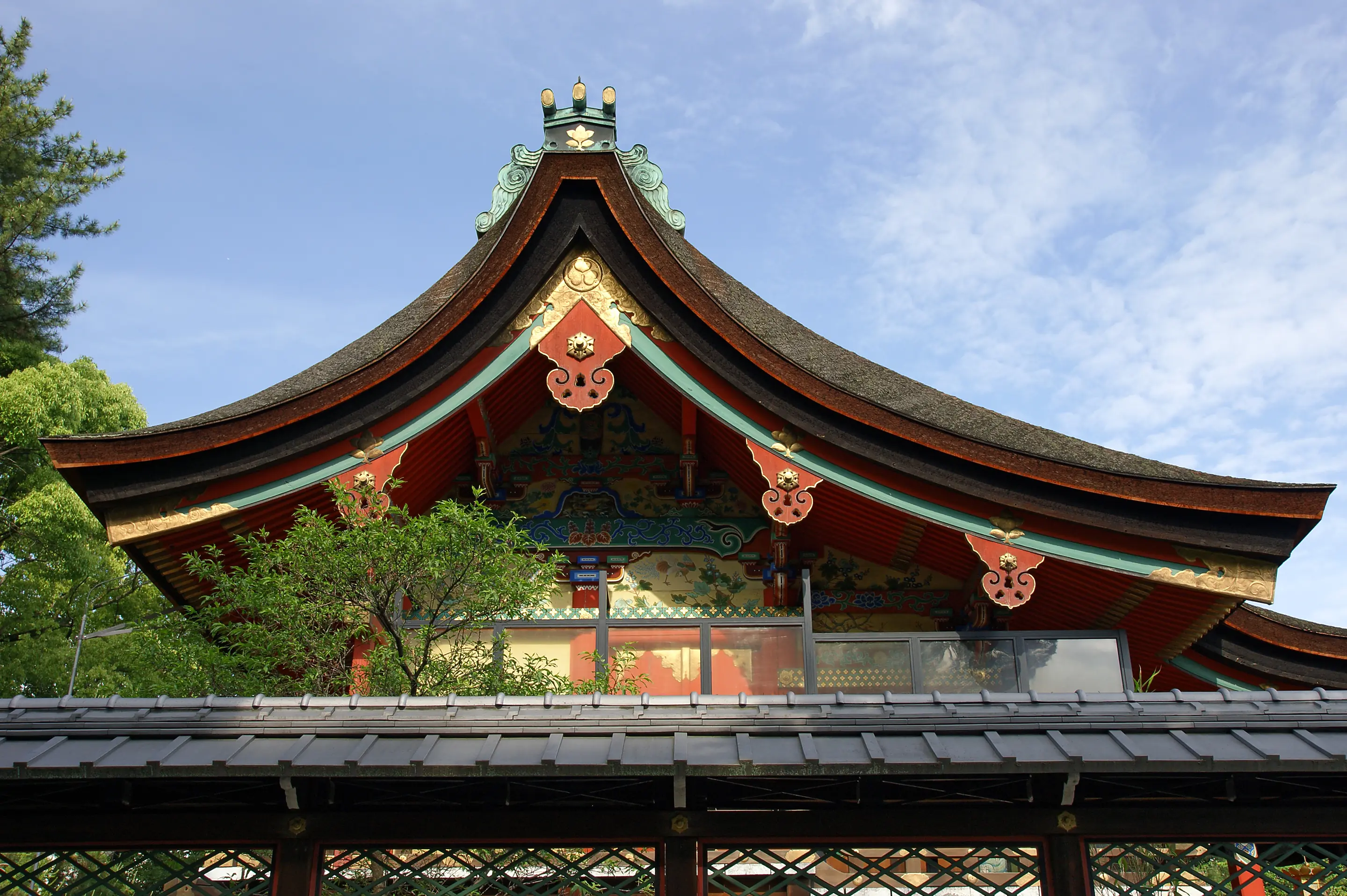 Curved shrine roof and gable with red, gold and green carvings under a blue sky, Gokōnomiya Shrine, Fushimi, Kyoto