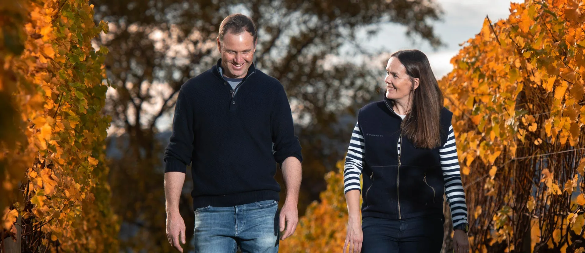 A man and a woman smile as they walk between vine rows in orange autumn leaf, at Te Whare Ra