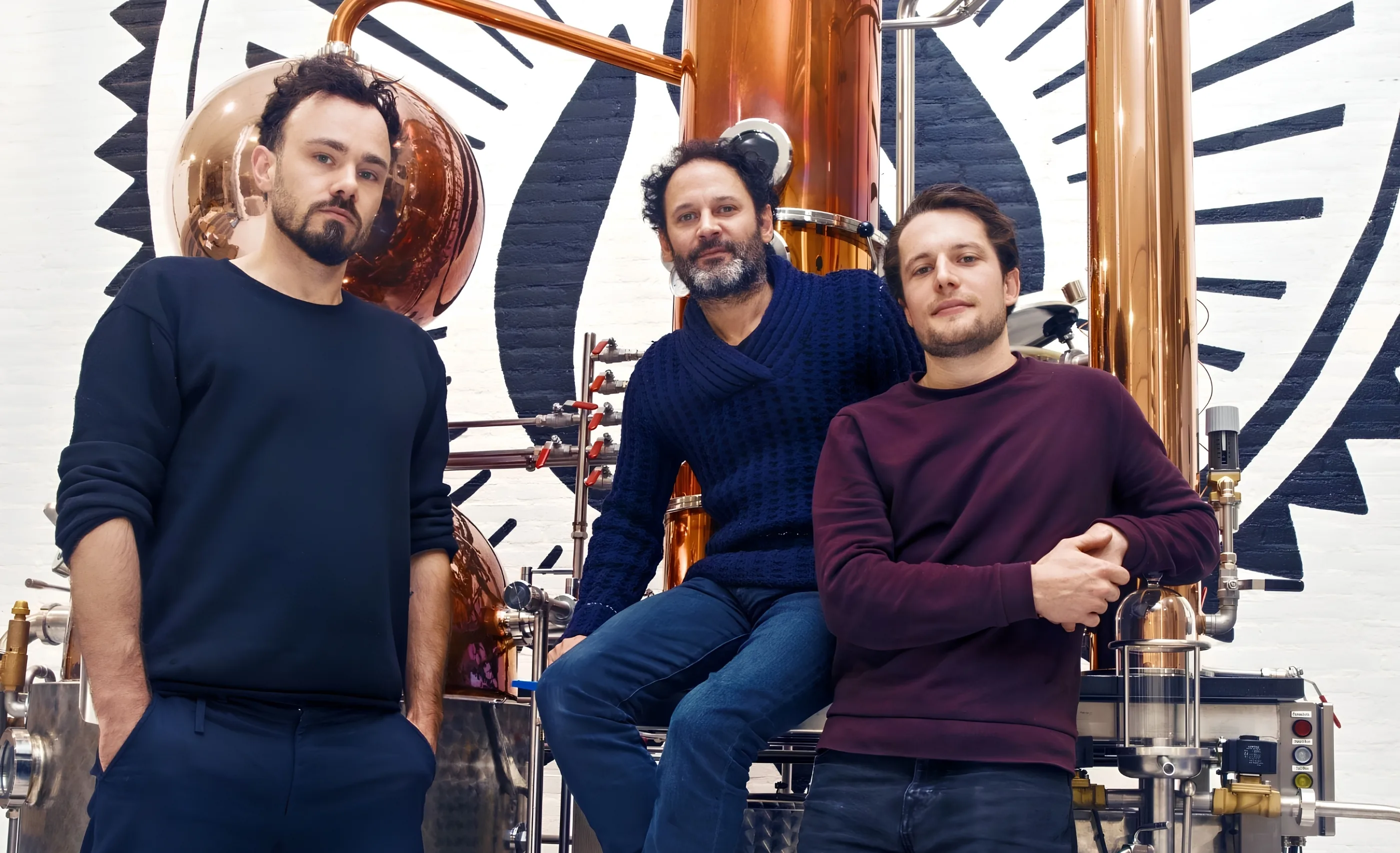 Three men in dark sweaters stand and sit before tall copper stills and a black-and-white painted sunburst mural