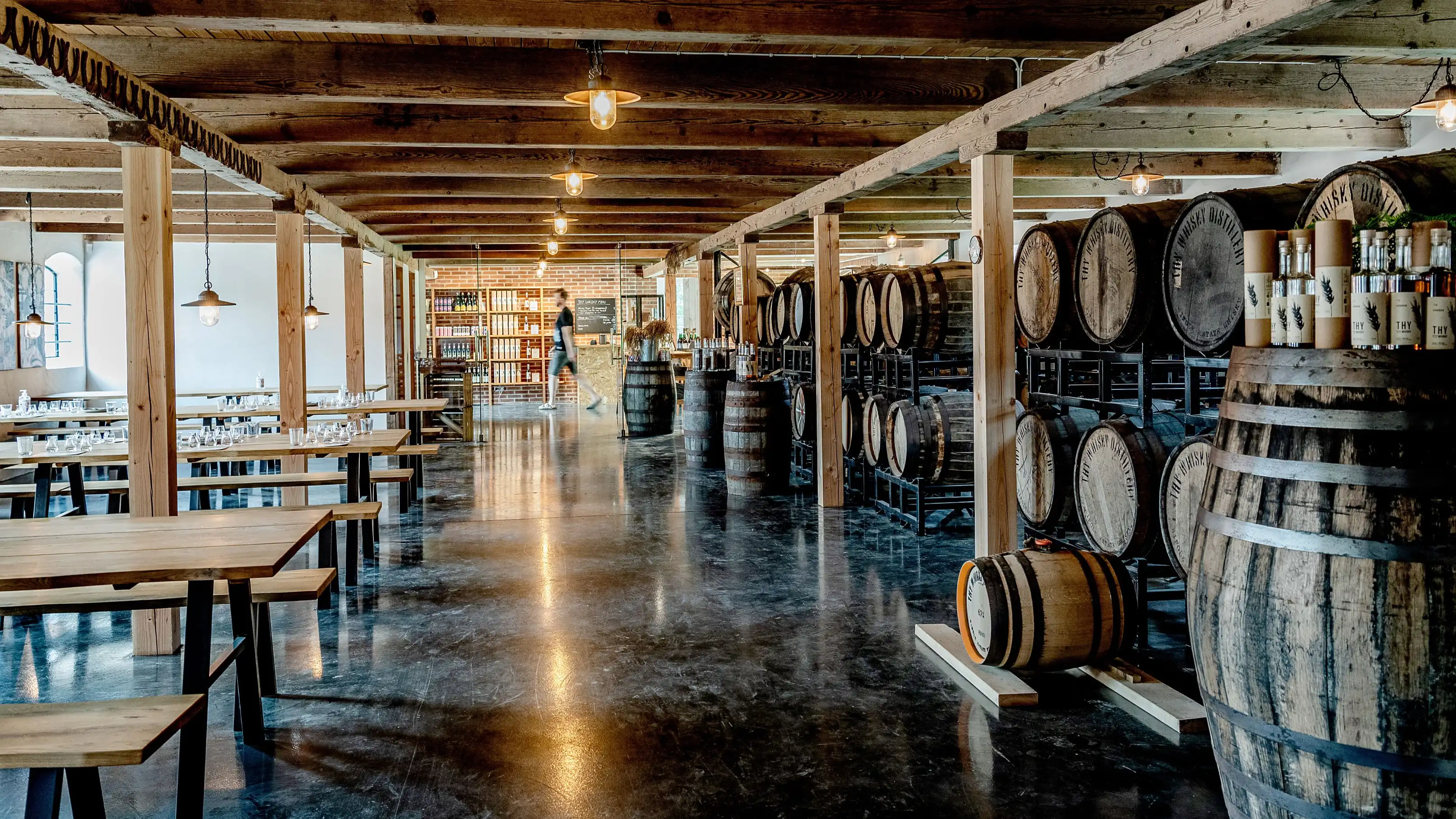 A timber-beamed hall with long wooden tables set with glasses, barrels on racks and shelves of bottles at the back, at Thy Whisky