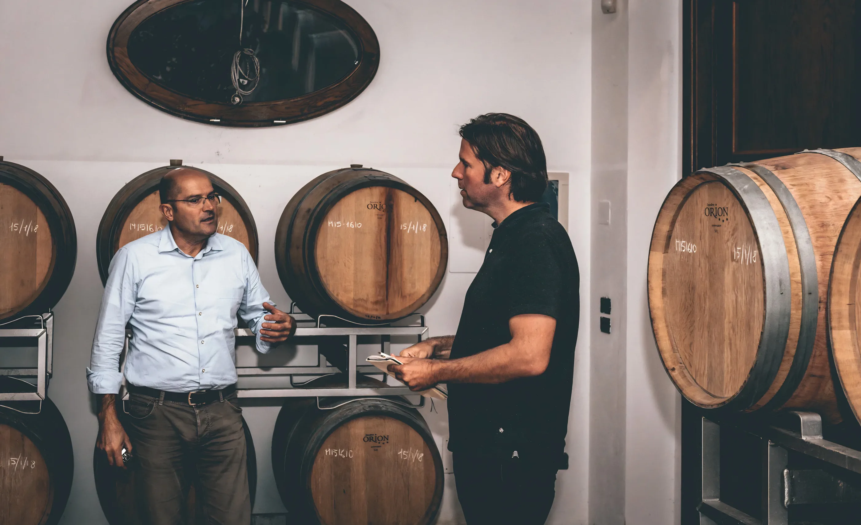 Two men talk in a cellar beside oak barrels chalked with dates, one in a pale blue shirt, one in black holding a notebook