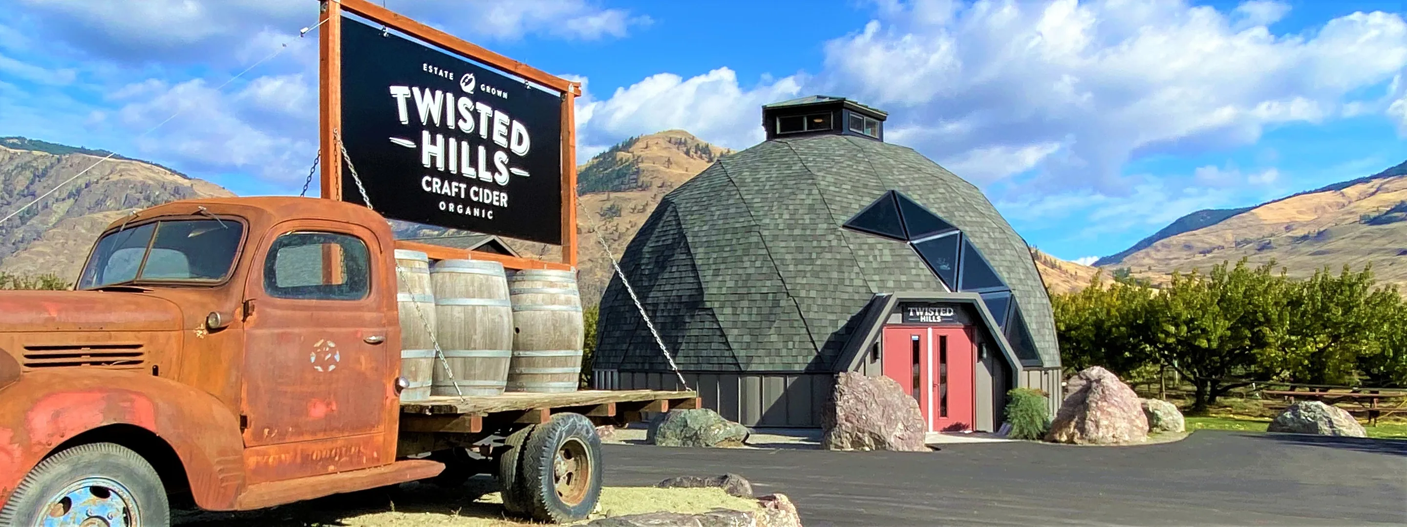 Geodesic dome with red doors, a rusty old truck carrying three barrels and a Twisted Hills sign, with orchard and dry hills